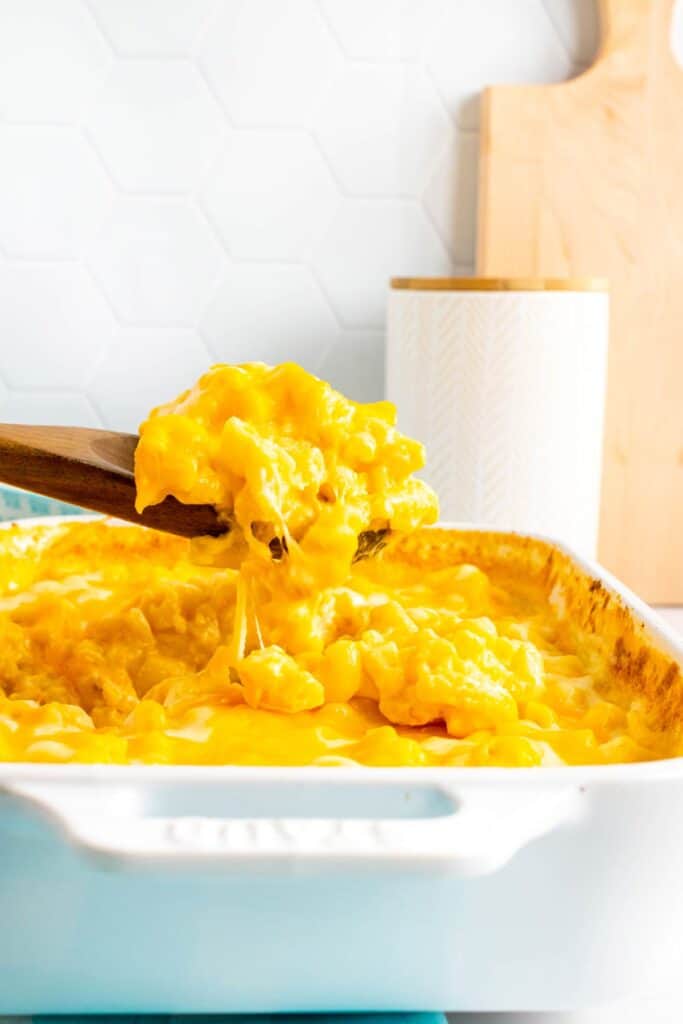 A side view of a baking dish filled with macaroni and cheese. A large wooden serving spoon is holding a large scoop of mac and cheese as melted cheese falls off the spoon.