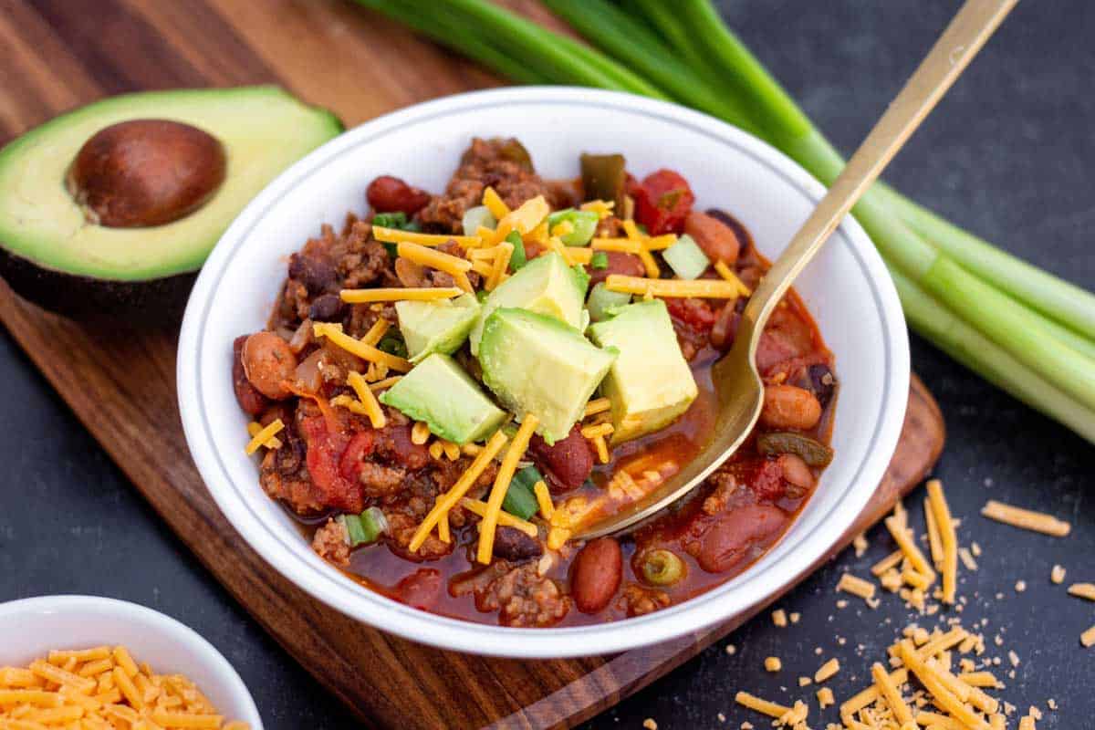 Small bowl of ground beef and sausage chili with shredded cheddar and diced avocado on top.
