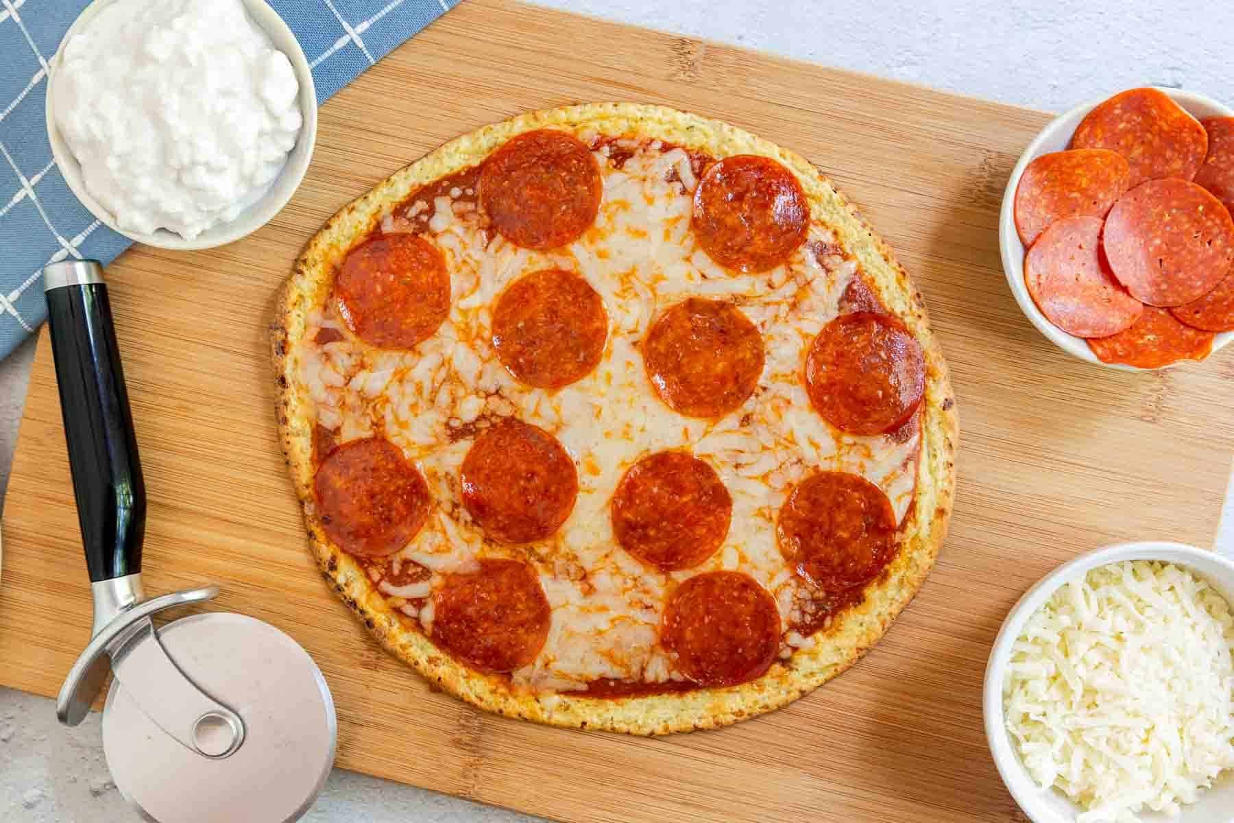 A whole pepperoni pizza on a wooden board with bowls of cottage cheese, shredded mozzarella, and pepperoni.