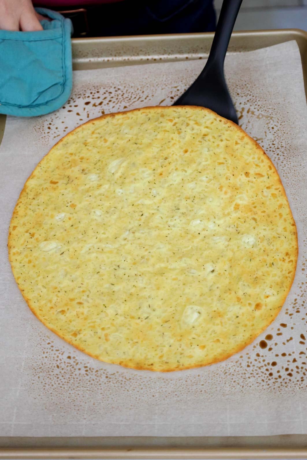 A baked pizza crust on a parchment-lined baking sheet being flipped with a spatula.