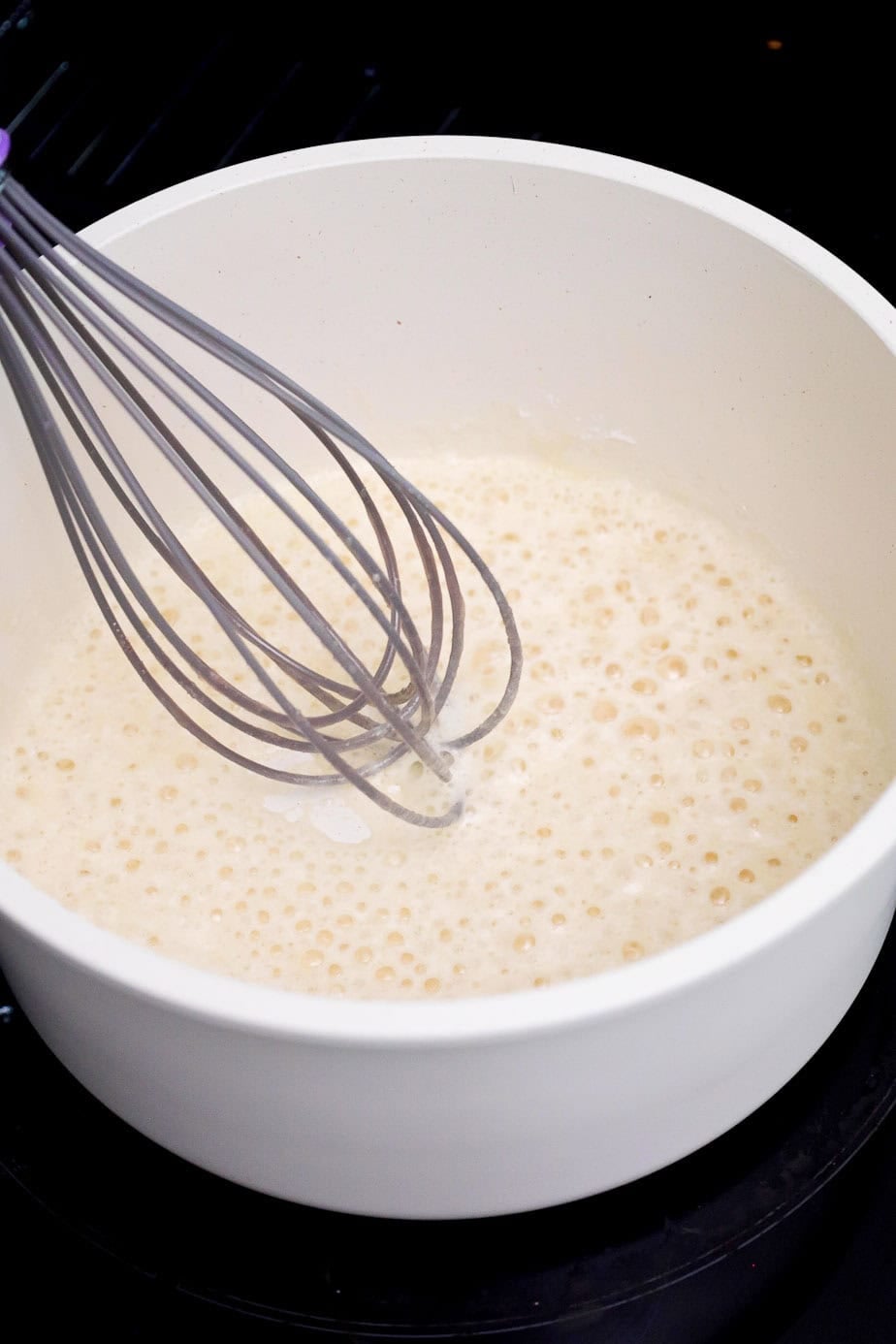 A whisk stirring bubbling melted butter and flour in a white pot.