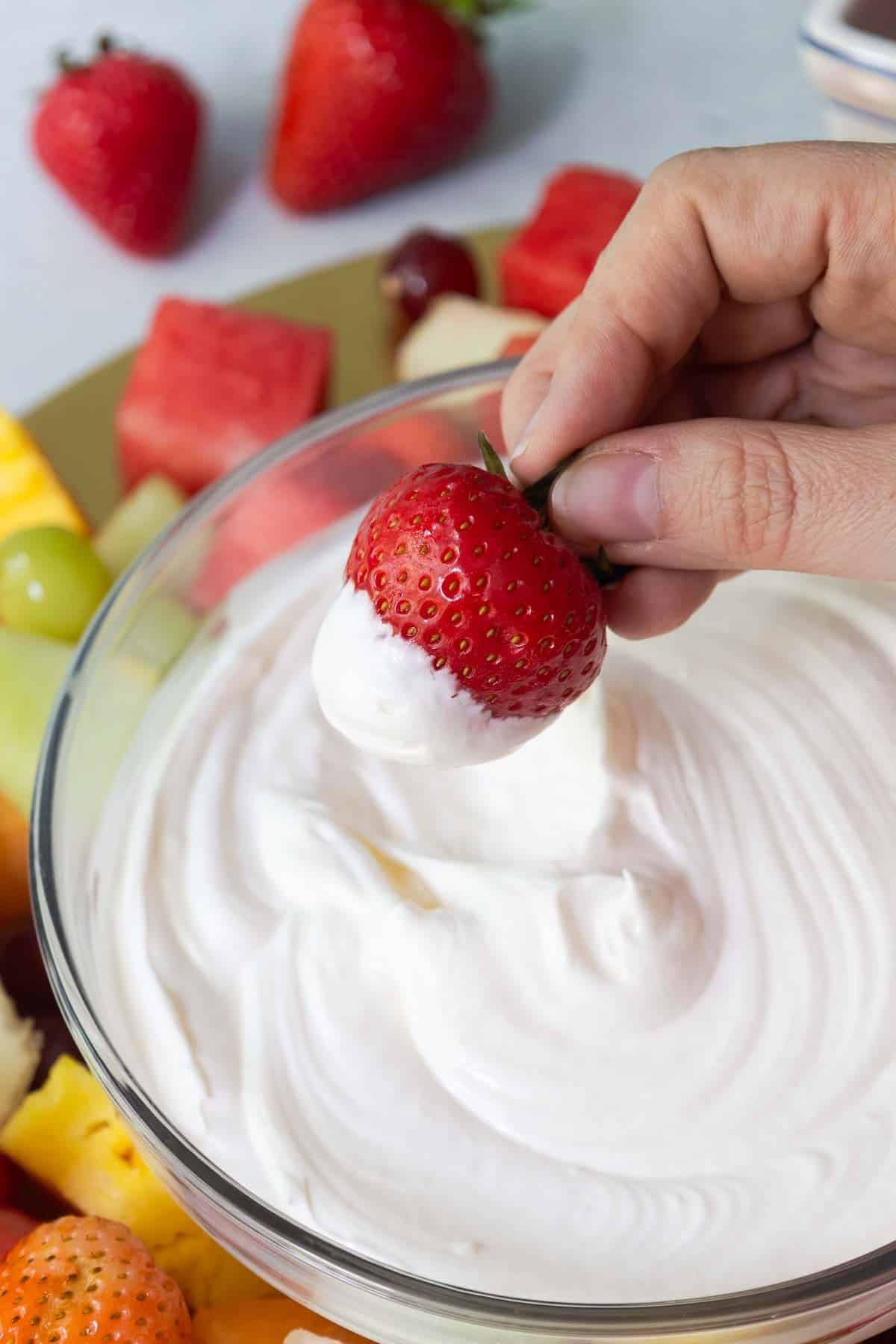A hand holding a fresh strawberry dipped halfway into a creamy white fruit dip, with a colorful fruit platter visible in the background.