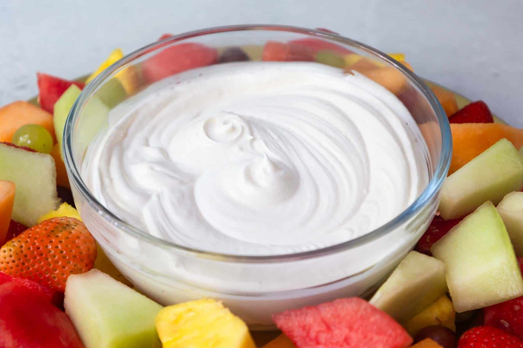A glass bowl of smooth, swirled white fruit dip in the center of a vibrant assortment of cut fruit pieces arranged on a platter.
