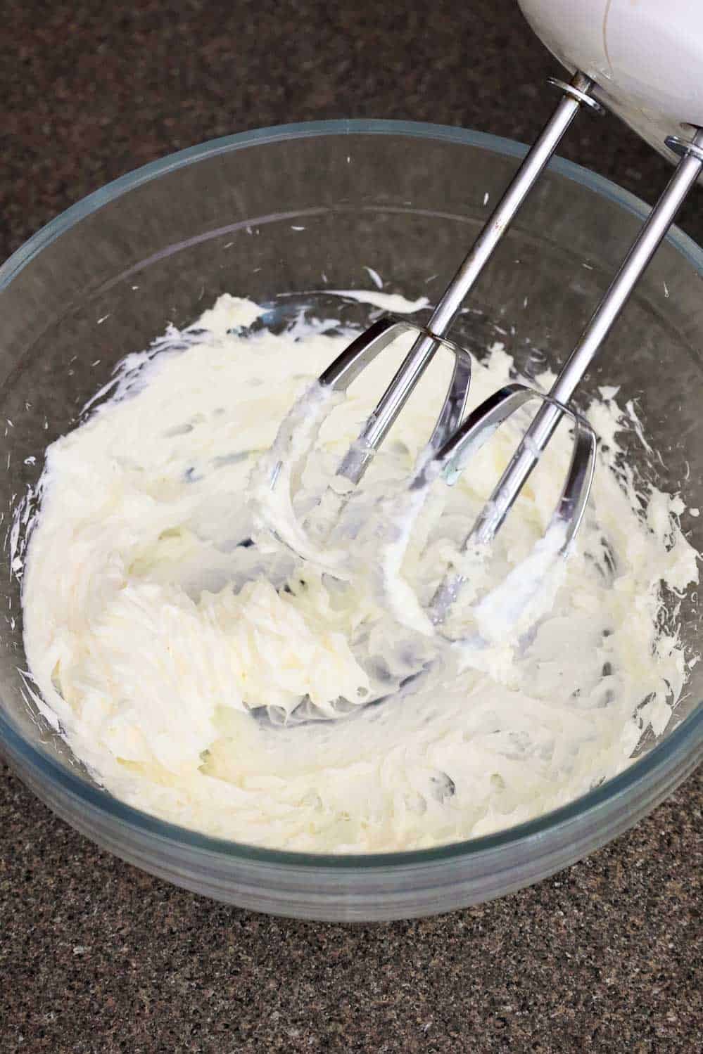 A bowl of cream cheese being mixed with an electric hand mixer.