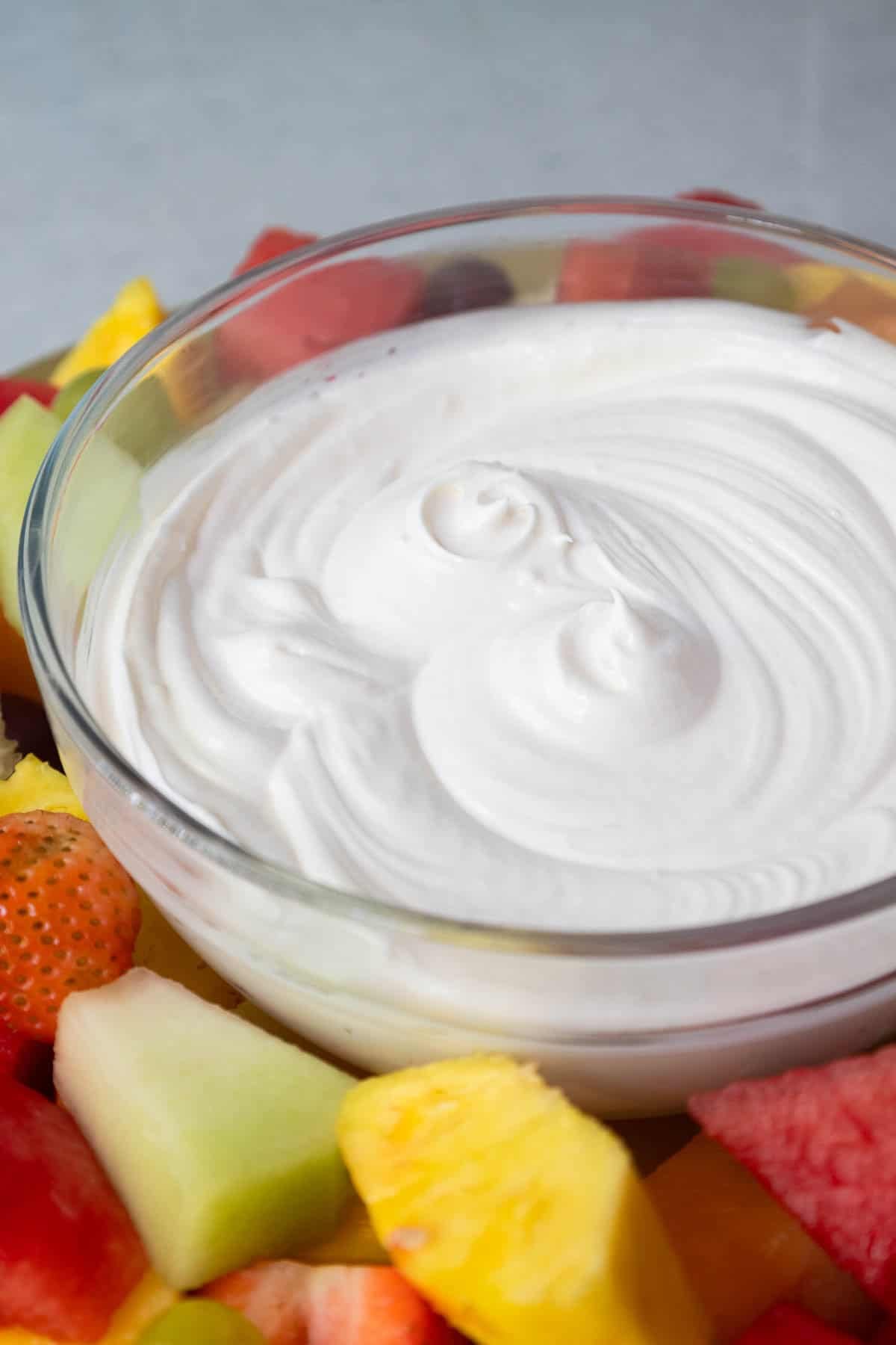 Close-up of a swirled bowl of white fruit dip surrounded by colorful chunks of fresh fruit.
