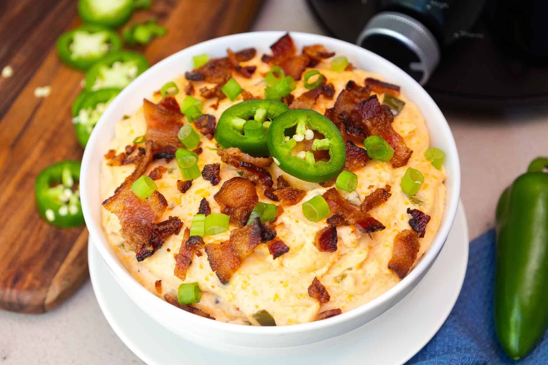 Close-up of jalapeño popper dip in a white bowl, garnished with chopped bacon, green onions, and fresh jalapeño slices, with a Crockpot in the background.