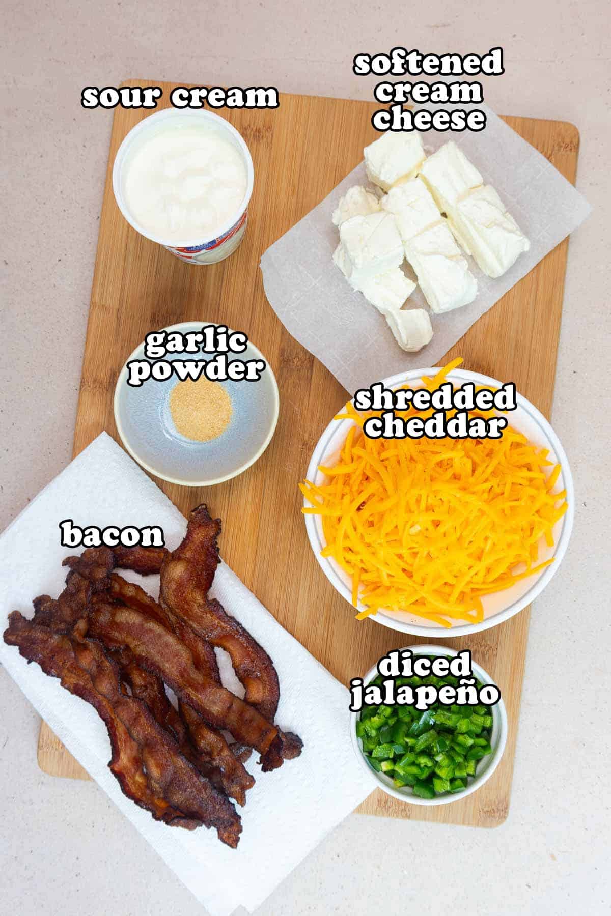 Overhead view of the main ingredients for crockpot jalapeño popper dip on a wooden board, including bacon, shredded cheddar, cream cheese, sour cream, diced jalapeños, and garlic powder, each labeled.