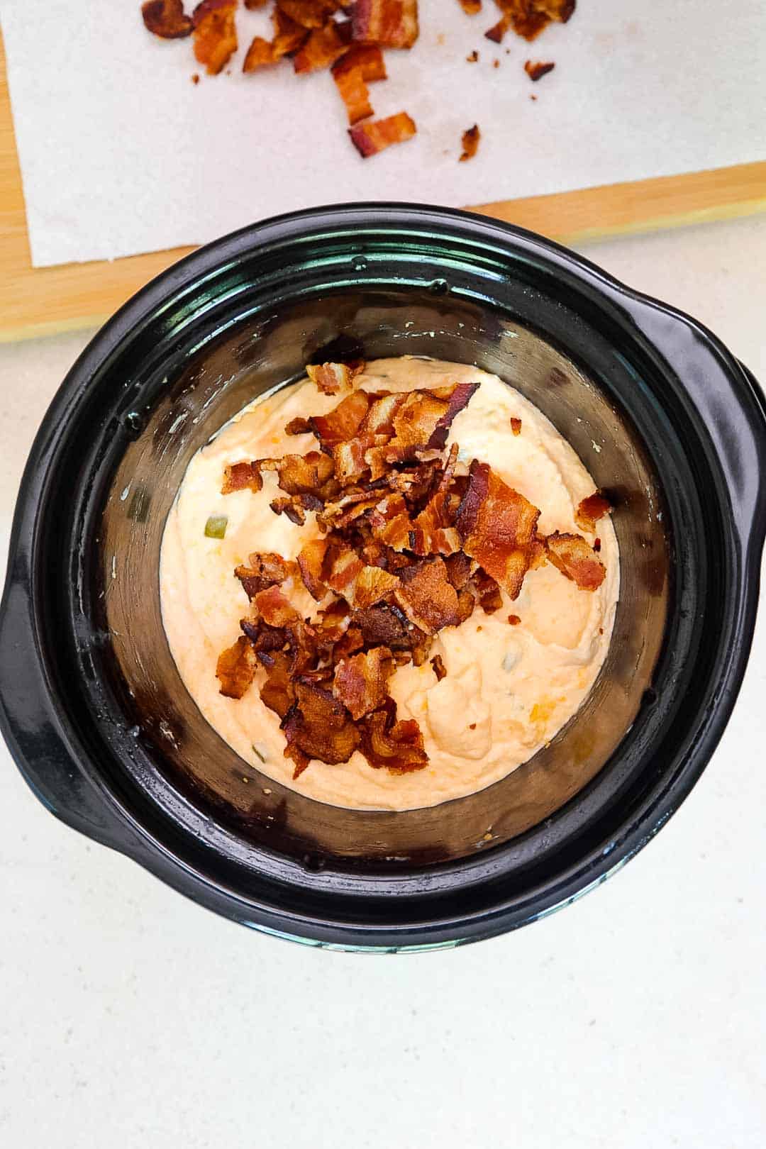 Slow cooker with cooked jalapeño popper dip topped with a generous amount of chopped bacon, before final mixing.