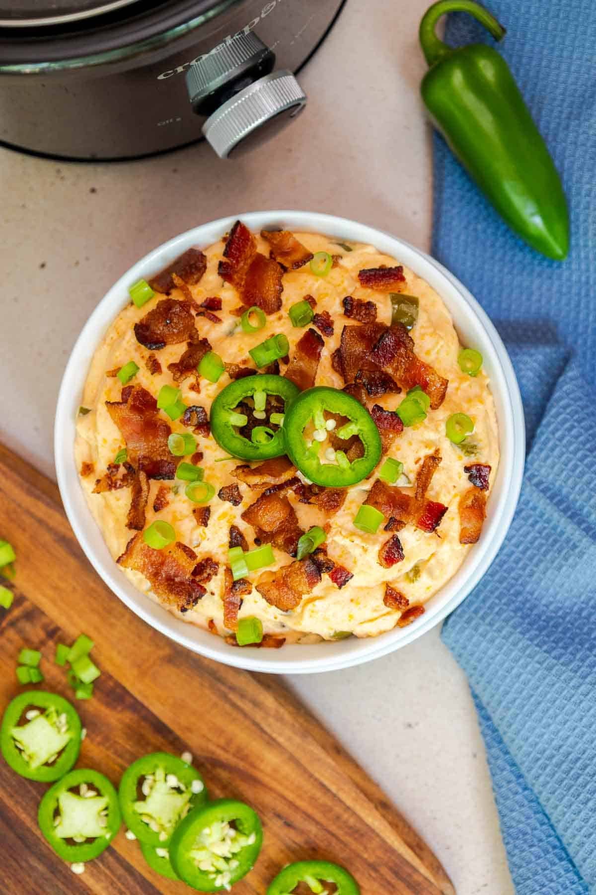 Overhead shot of finished crockpot jalapeño popper dip in a white bowl, garnished with sliced jalapeños, crispy bacon, and green onions, next to a green jalapeño and a Crockpot.