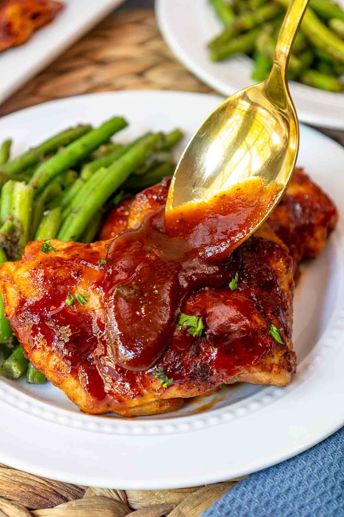 Golden spoon drizzling extra BBQ sauce over a serving of baked boneless skinless chicken thighs.