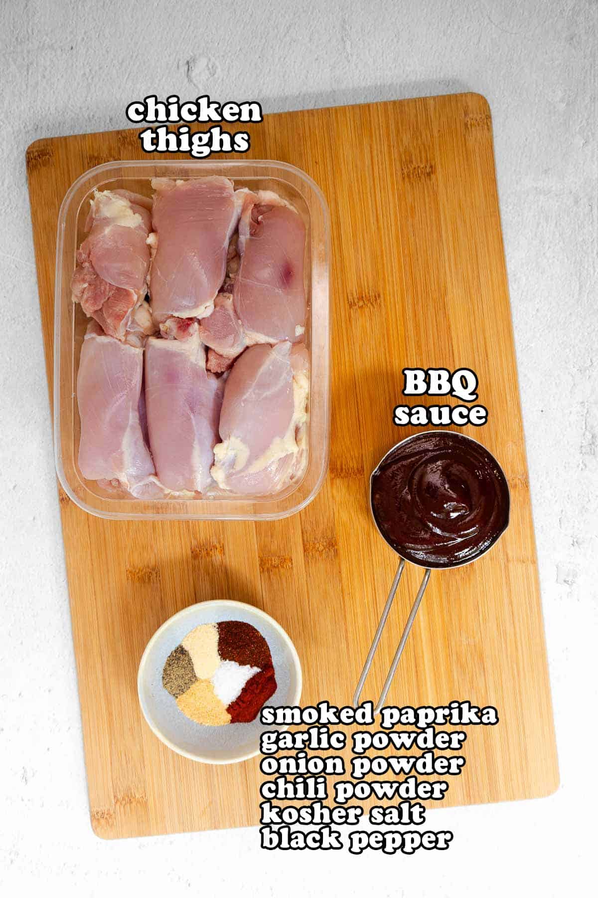 Flat lay of raw ingredients on a wooden cutting board including boneless chicken thighs, a bowl of BBQ sauce, and a spice blend labeled with smoked paprika, garlic powder, onion powder, chili powder, kosher salt, and black pepper.