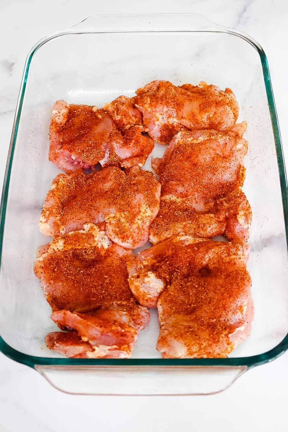Raw boneless chicken thighs evenly coated in dry spice rub, arranged in a glass baking dish before cooking.