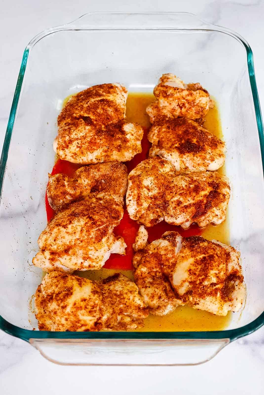 Seasoned chicken thighs placed in a glass dish with liquid pooling underneath, ready for the oven.