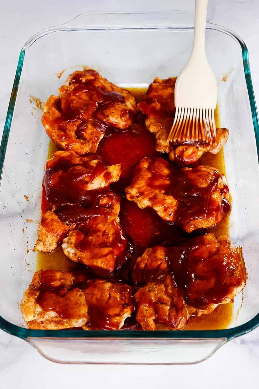 Chicken thighs being brushed with BBQ sauce using a silicone brush, midway through baking in a glass dish.