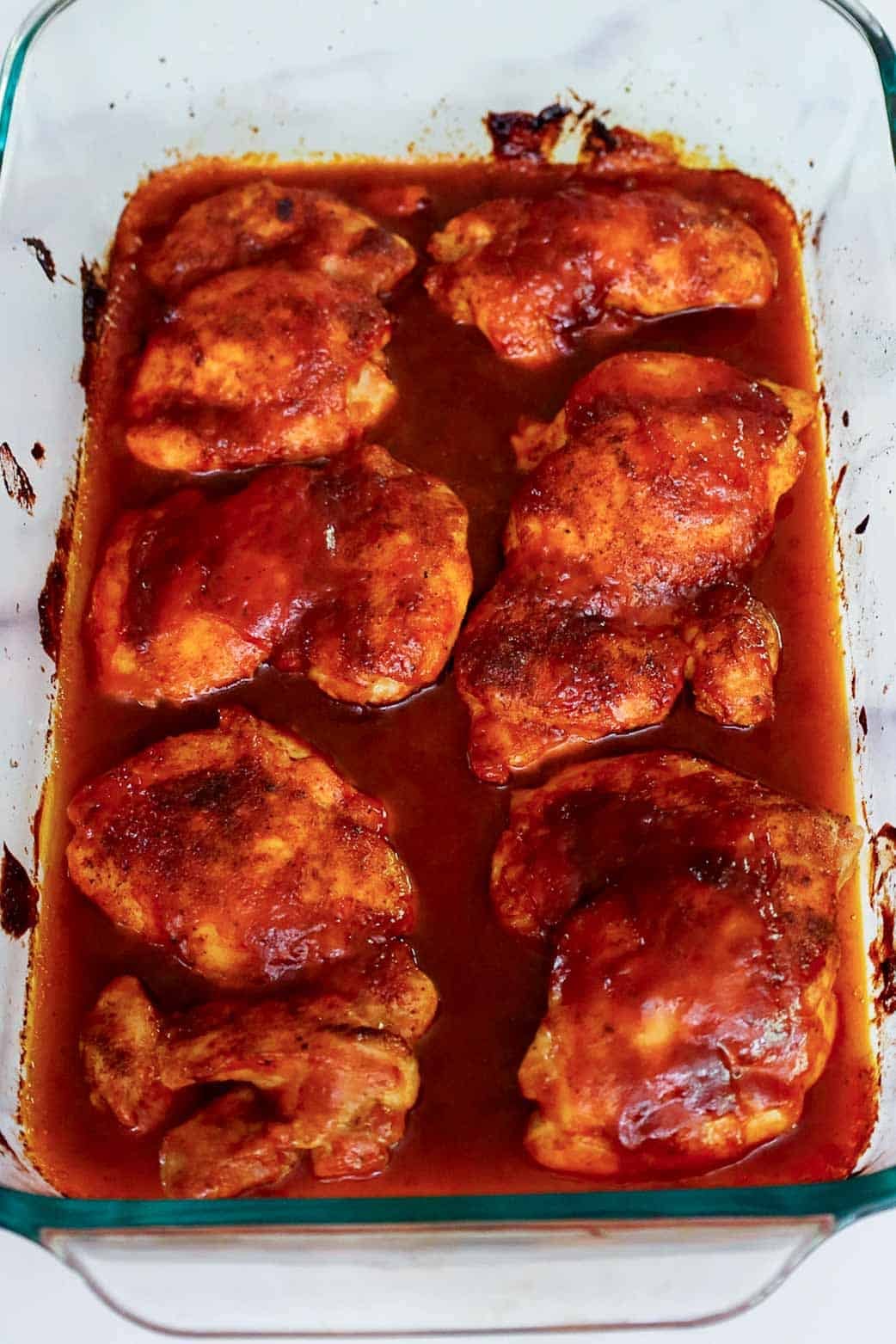 Fully cooked oven baked BBQ boneless chicken thighs resting in a glass baking dish, coated in a thick layer of caramelized sauce.