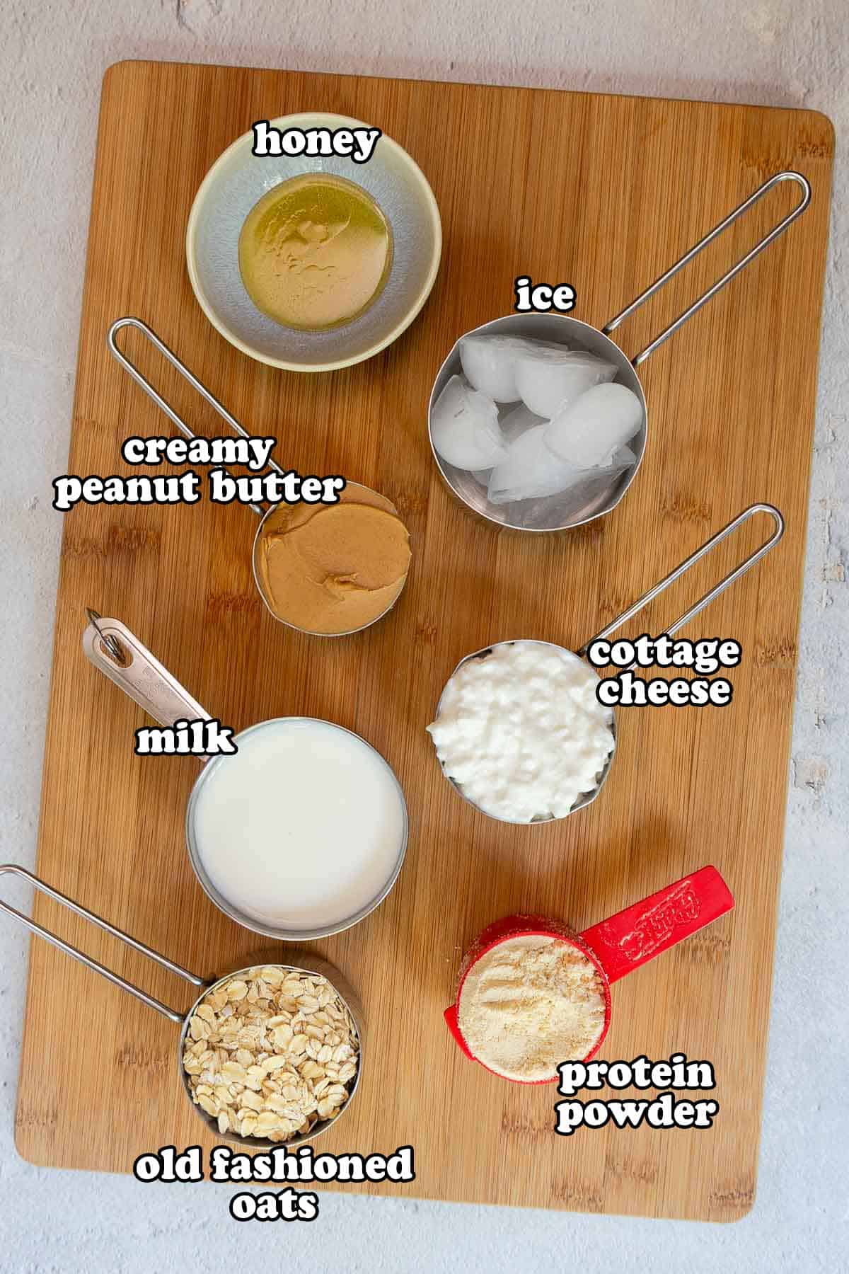 A wooden cutting board topped with smoothie ingredients labeled: honey, ice, creamy peanut butter, milk, old fashioned oats, cottage cheese, and protein powder.