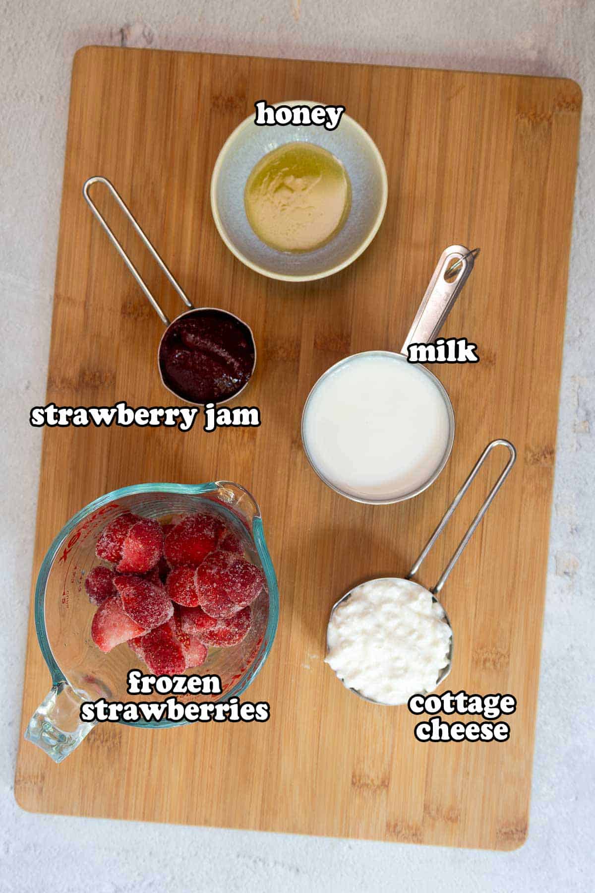 Ingredients for the jelly smoothie on a wooden cutting board, labeled: honey, milk, strawberry jam, frozen strawberries, and cottage cheese.