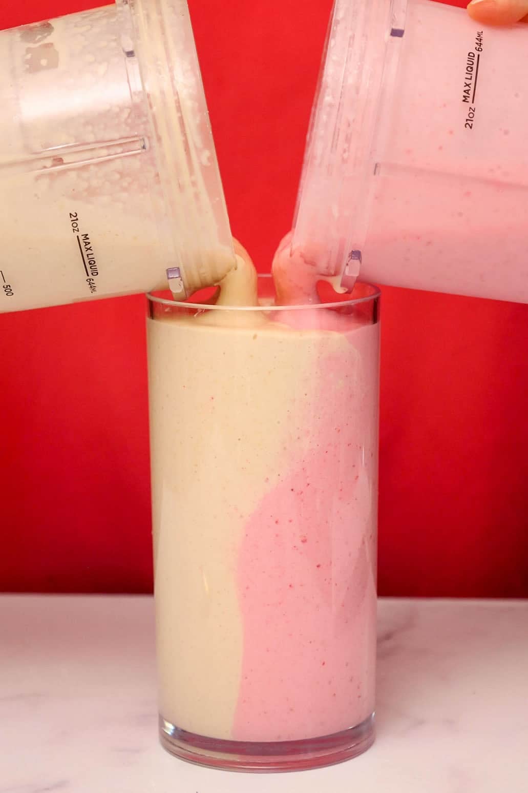 Two blender cups simultaneously pouring the peanut butter and jelly smoothies side-by-side into a single glass, creating a swirled effect.
