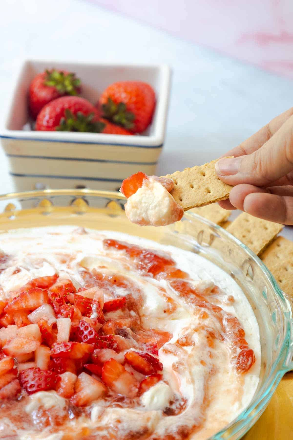 Close-up of a hand dipping a graham cracker into a creamy strawberry dessert, topped with fresh strawberry chunks.
