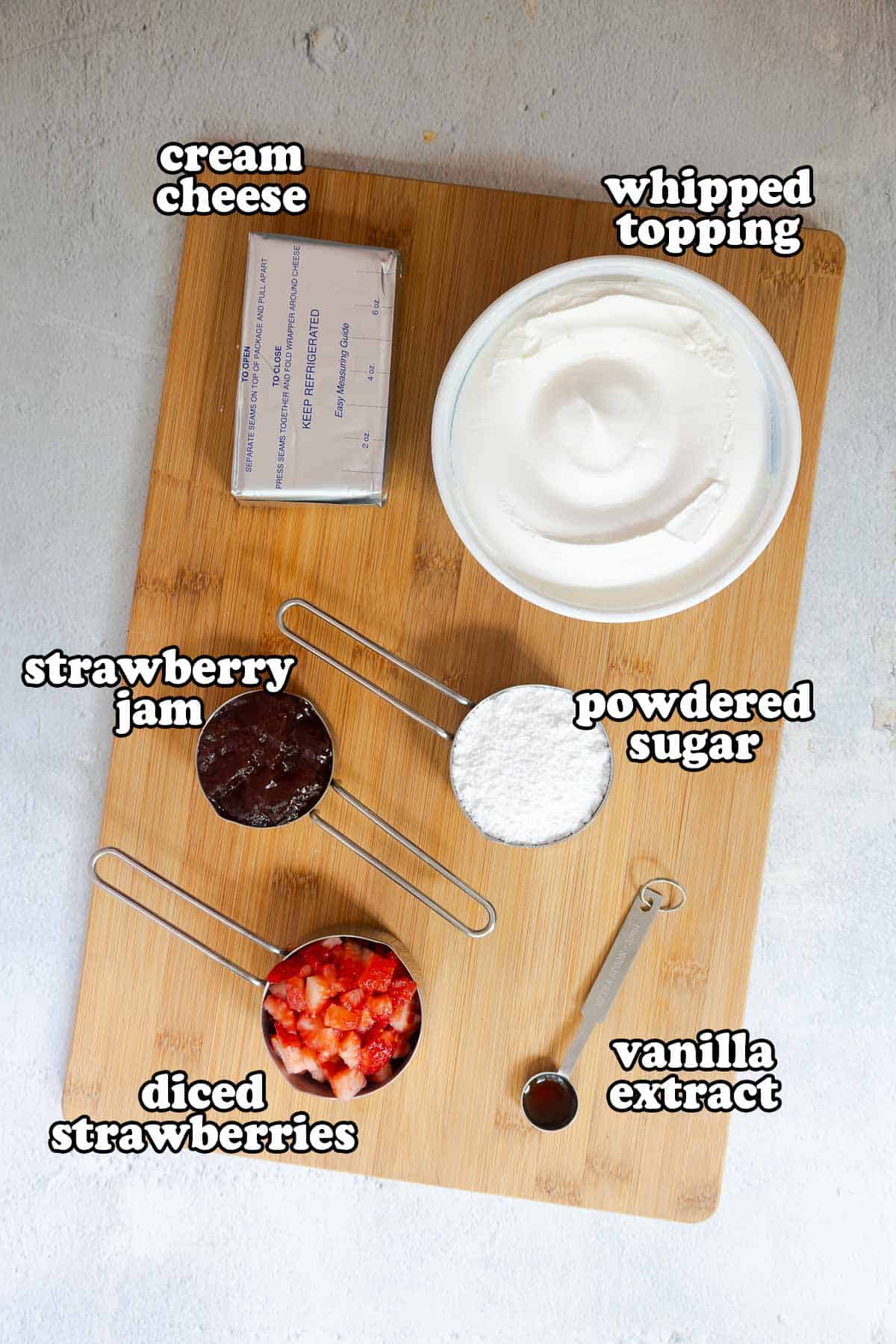 Ingredients for strawberry cheesecake dip on a wooden board, labeled as cream cheese, whipped topping, powdered sugar, strawberry jam, diced strawberries, and vanilla extract.