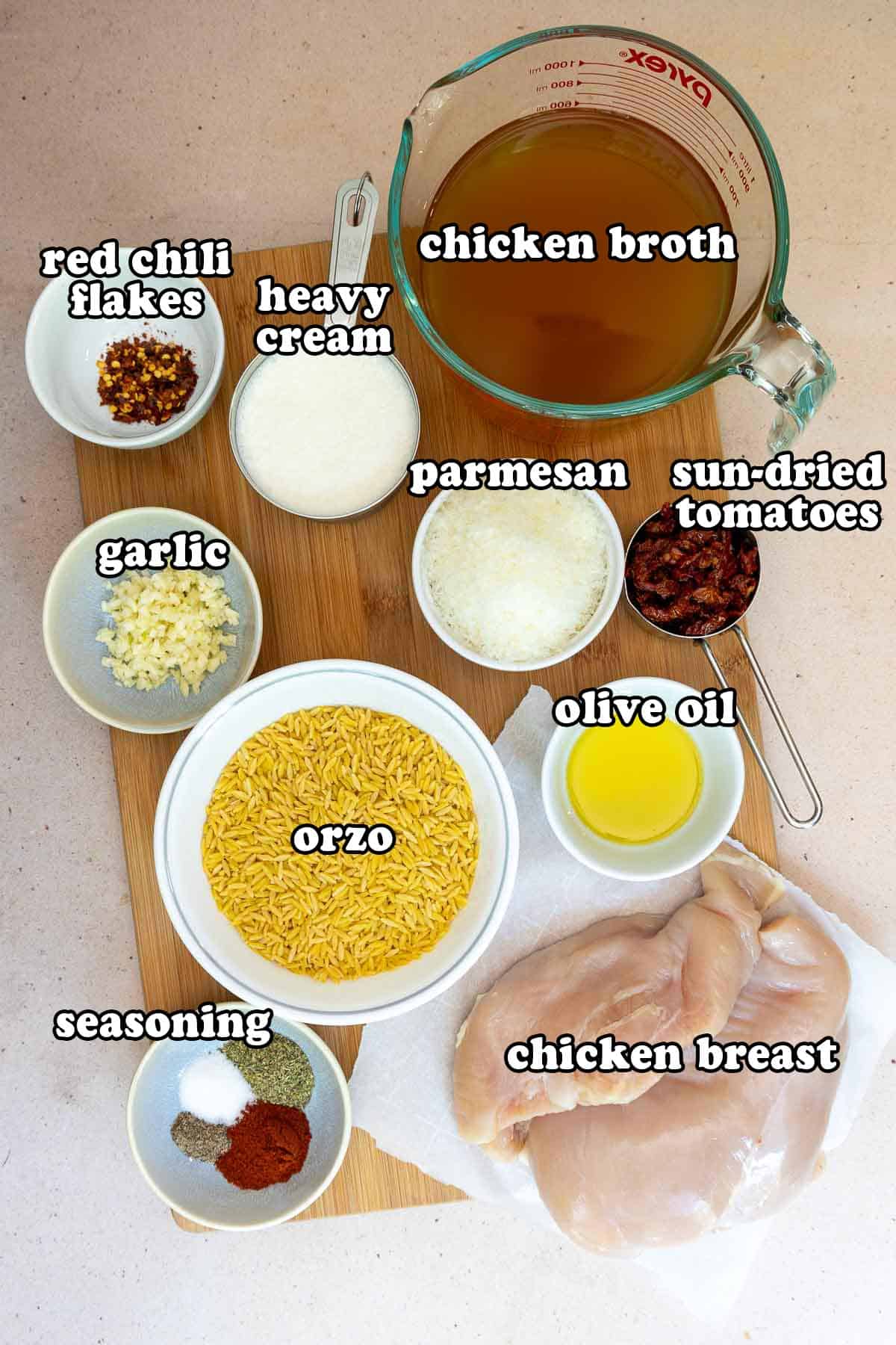 Flat lay of ingredients for creamy chicken orzo, including chicken broth, sun-dried tomatoes, olive oil, raw chicken breasts, orzo, garlic, parmesan, heavy cream, red chili flakes, and a bowl of seasoning spices.