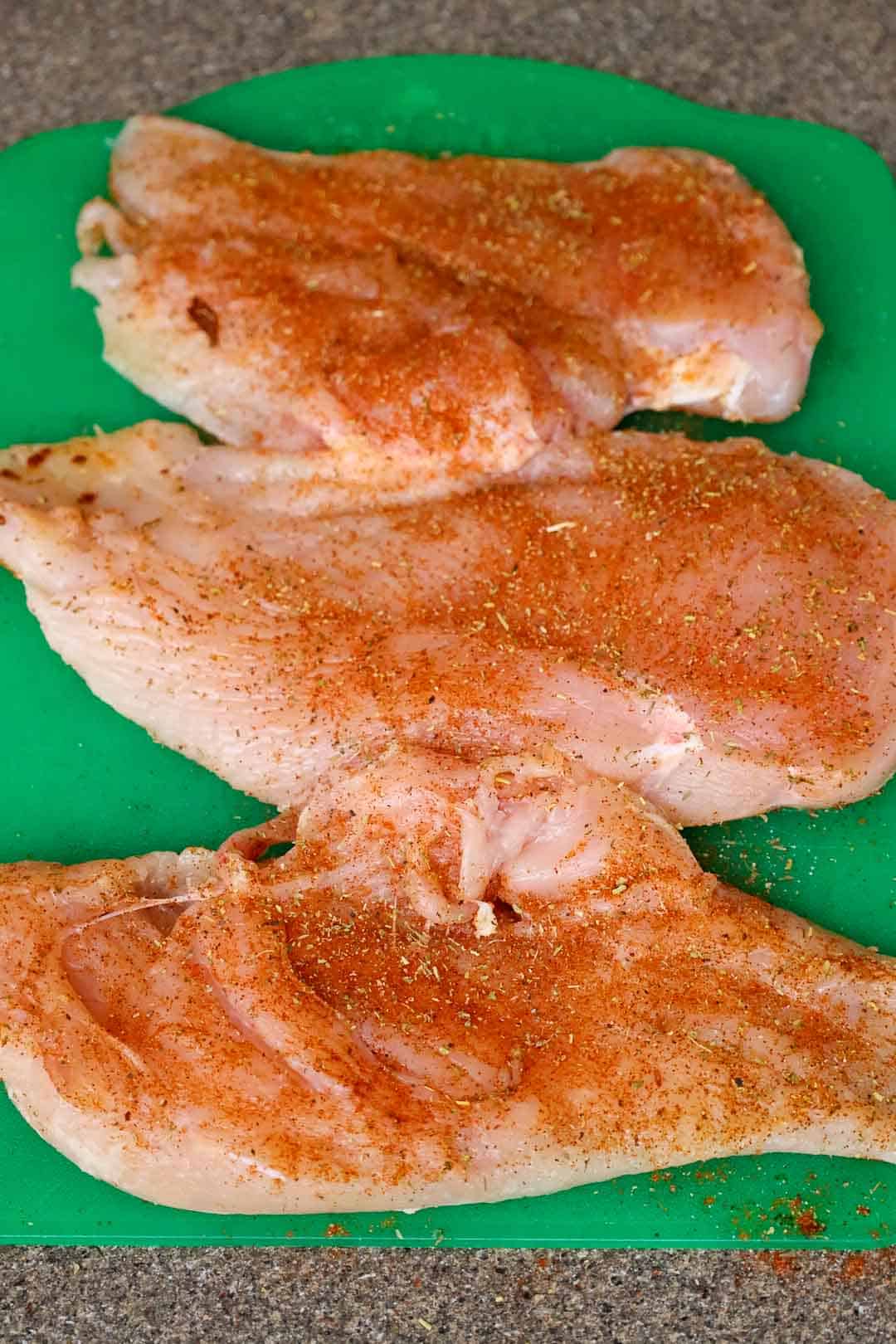 Three halved chicken breasts coated with seasoning, laid out on a cutting board before cooking.