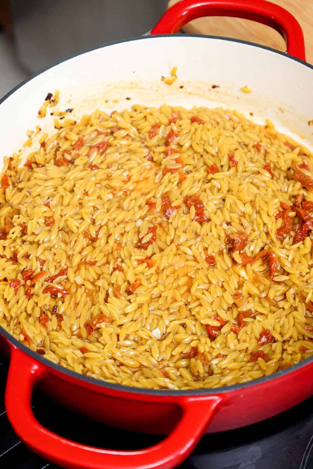 Orzo and sun-dried tomatoes cooking in chicken broth inside a Dutch oven, before cream and cheese are added.