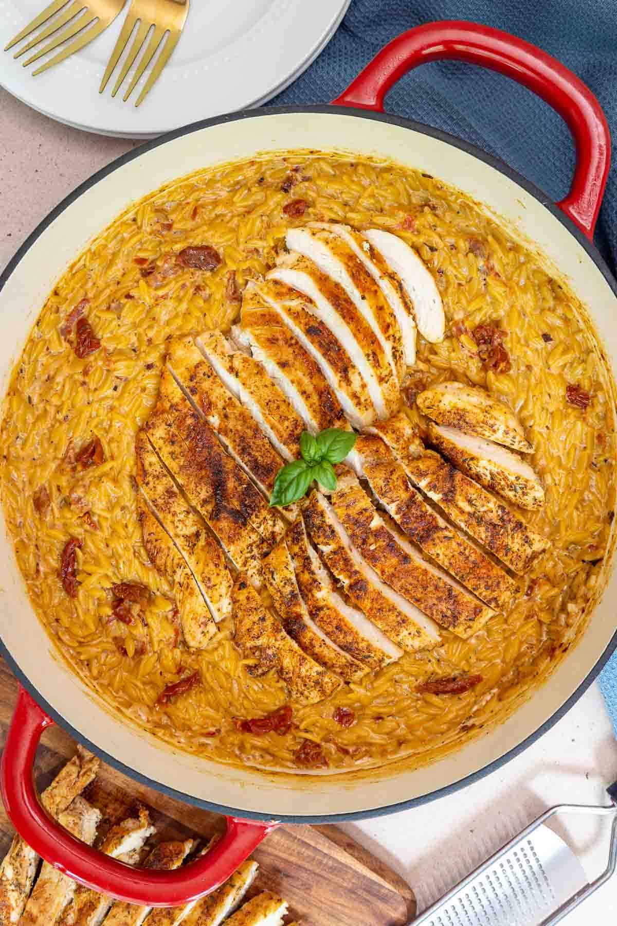Top-down view of a large pot of creamy sun-dried tomato orzo topped with sliced seasoned chicken breast and a basil garnish.