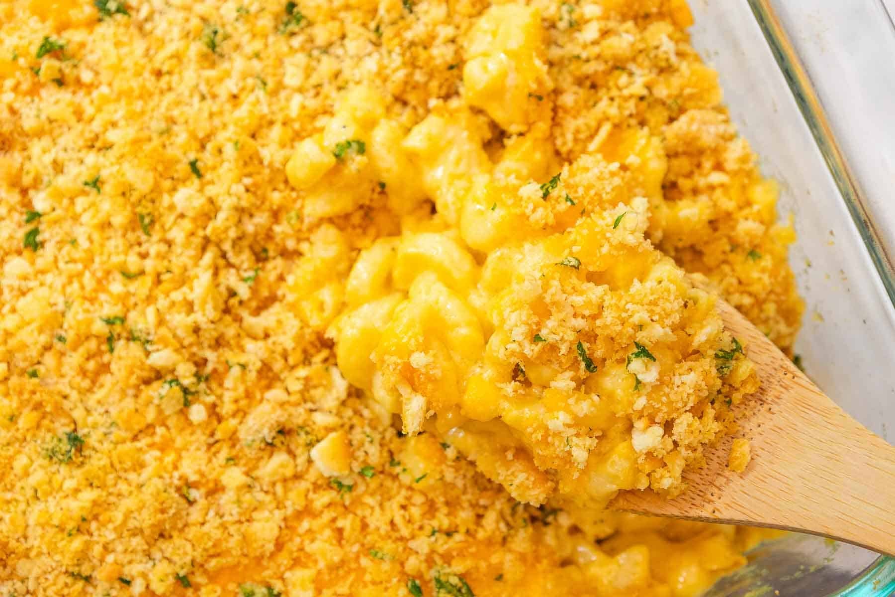 Wooden spoon scooping creamy cheddar mac and cheese topped with crumbled cracker topping out of a casserole dish.