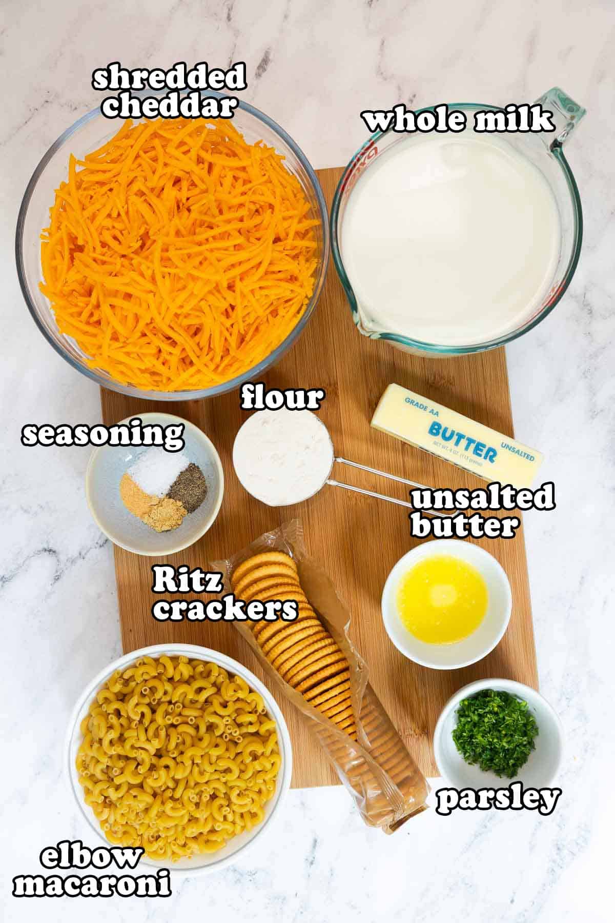 Ingredients for old fashioned baked mac and cheese with Ritz crackers displayed on a wooden board, including shredded cheddar, whole milk, unsalted butter, flour, elbow macaroni, Ritz crackers, seasoning, and parsley.