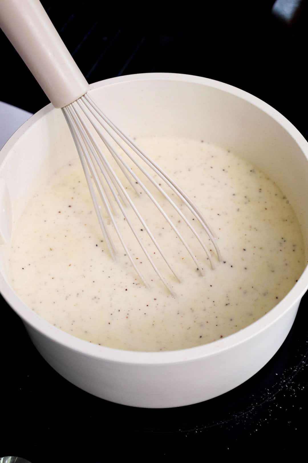 Flour and seasoning whisked into milk to create a béchamel-style base in a white saucepan during the early stages of making mac and cheese sauce.