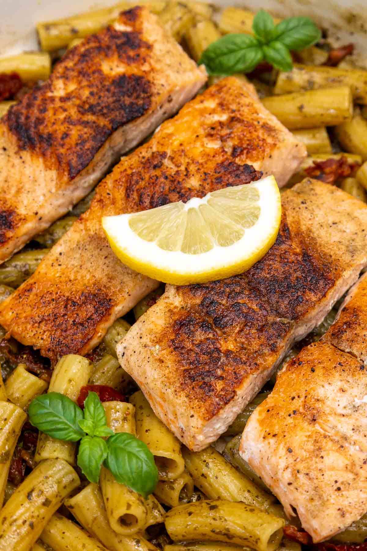 Four blackened salmon fillets arranged over pesto-coated pasta with sun-dried tomatoes and fresh basil