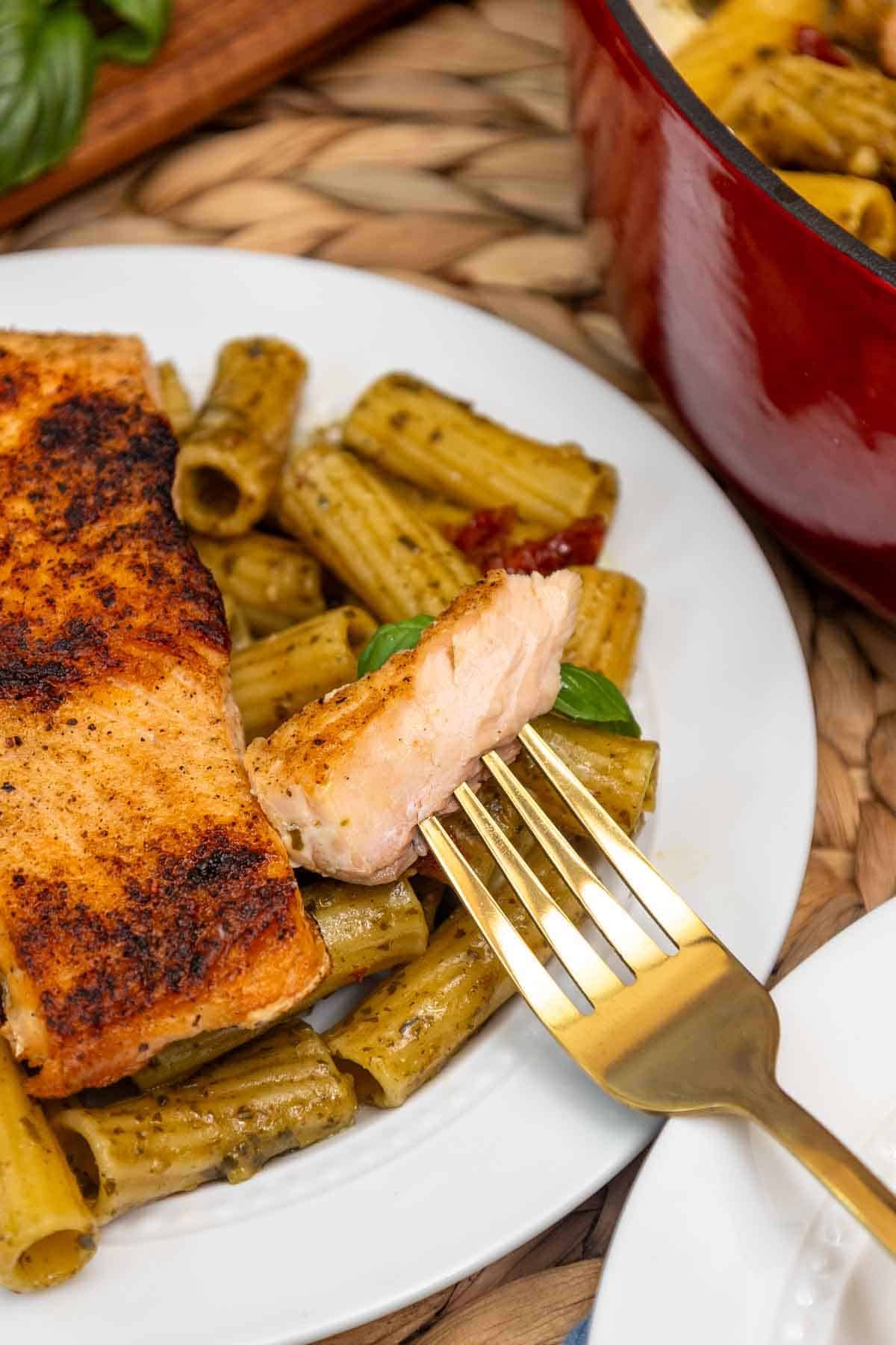 A fork lifting a bite-sized piece of flaky salmon from a plate of salmon pesto pasta, revealing the tender texture of the fish atop rigatoni coated in pesto sauce.