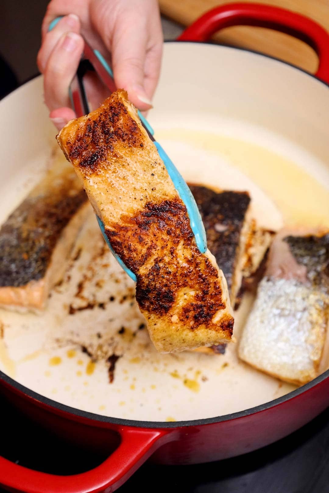 Close-up of a seared salmon fillet being lifted with tongs, showing a golden, crispy crust formed.