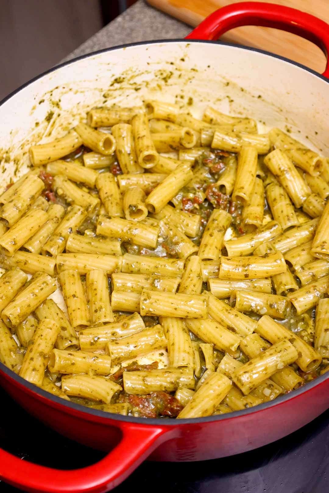 Cooked rigatoni pasta in a Dutch oven, coated in pesto sauce and mixed with bits of sun-dried tomatoes.
