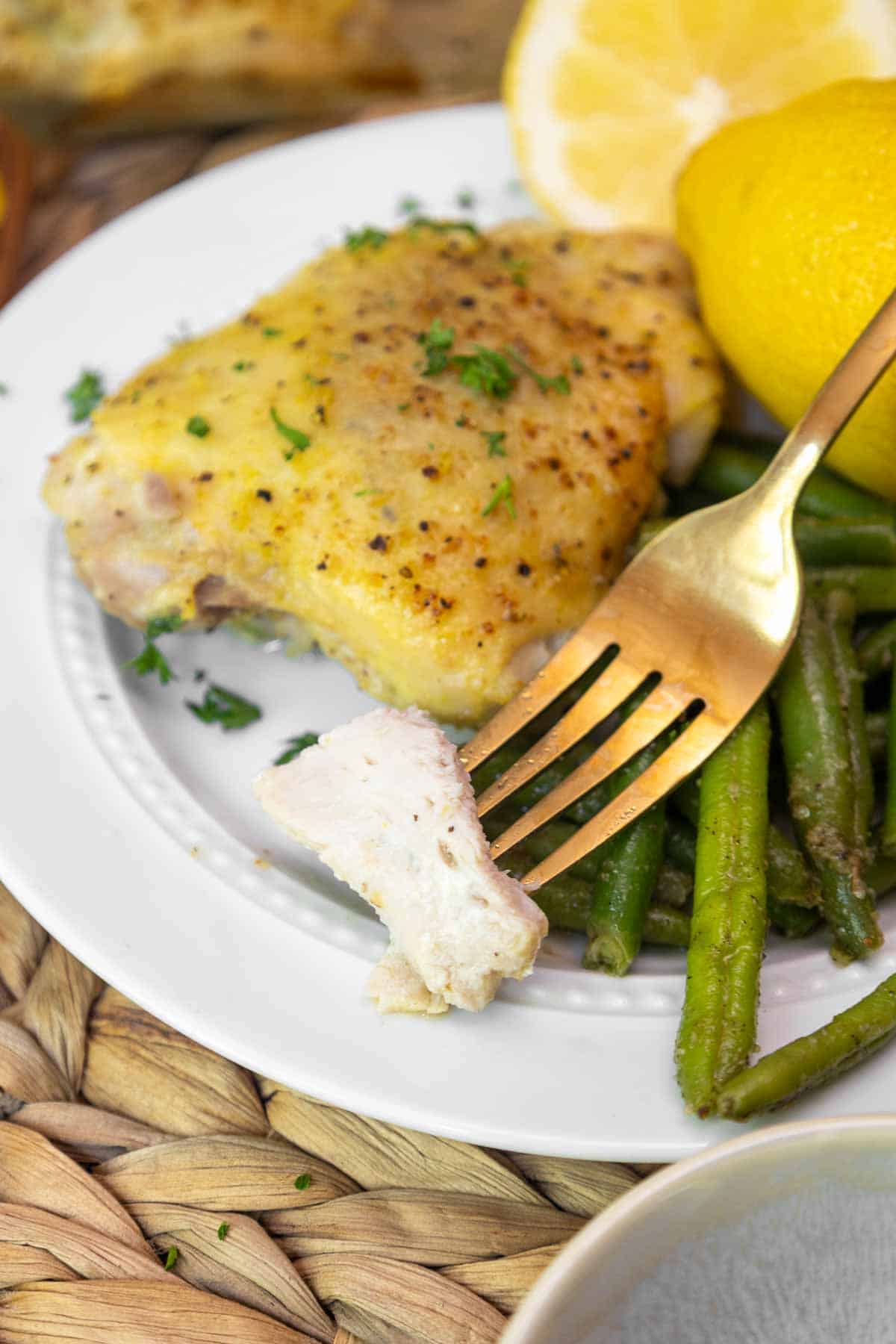 A fork holding a bite-sized piece of juicy baked chicken thigh above a plate of lemon pepper chicken, green beans, and fresh lemon.