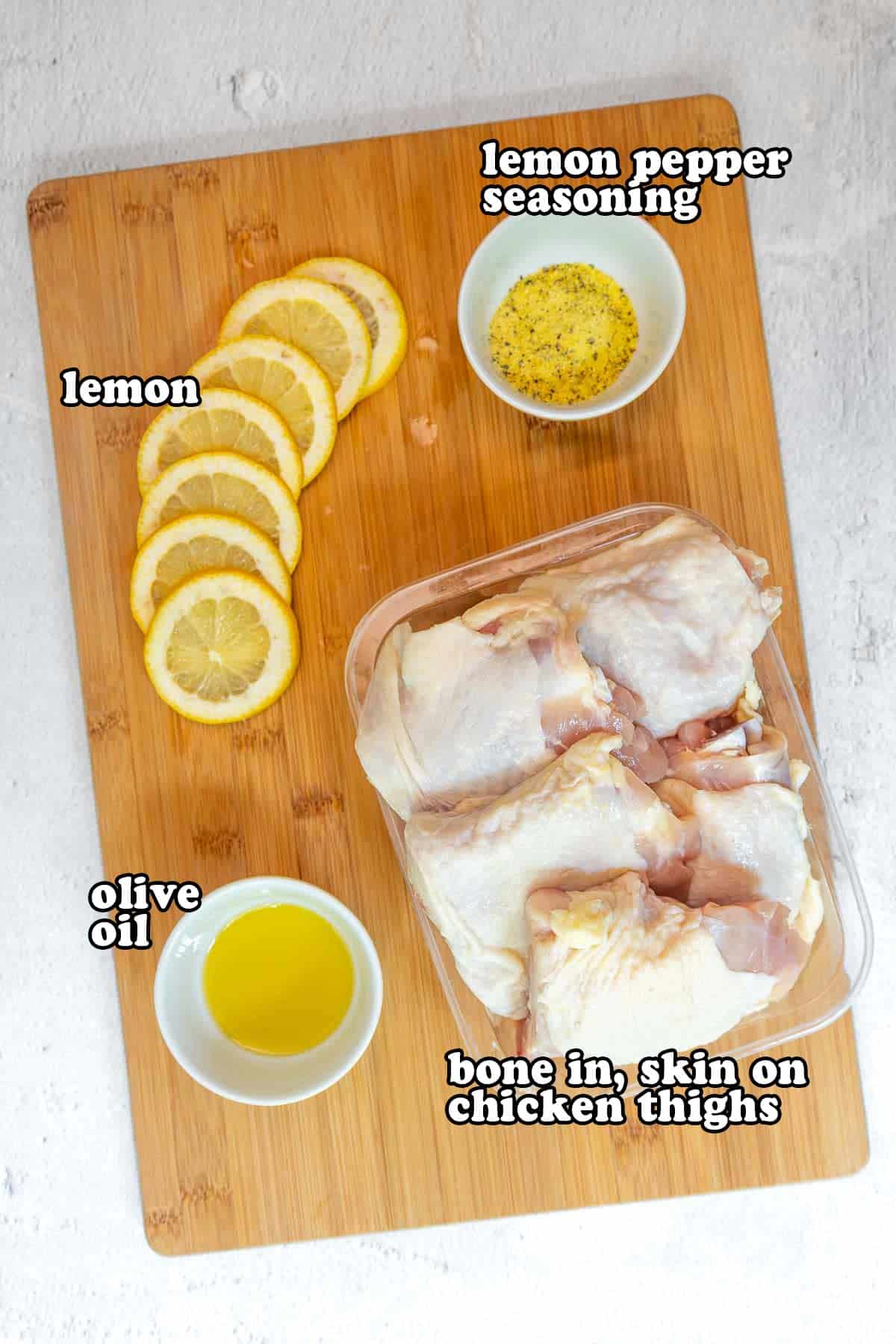 Ingredients for lemon pepper chicken laid out on a wooden cutting board, including sliced lemons, lemon pepper seasoning, olive oil, and raw chicken thighs with skin and bone.