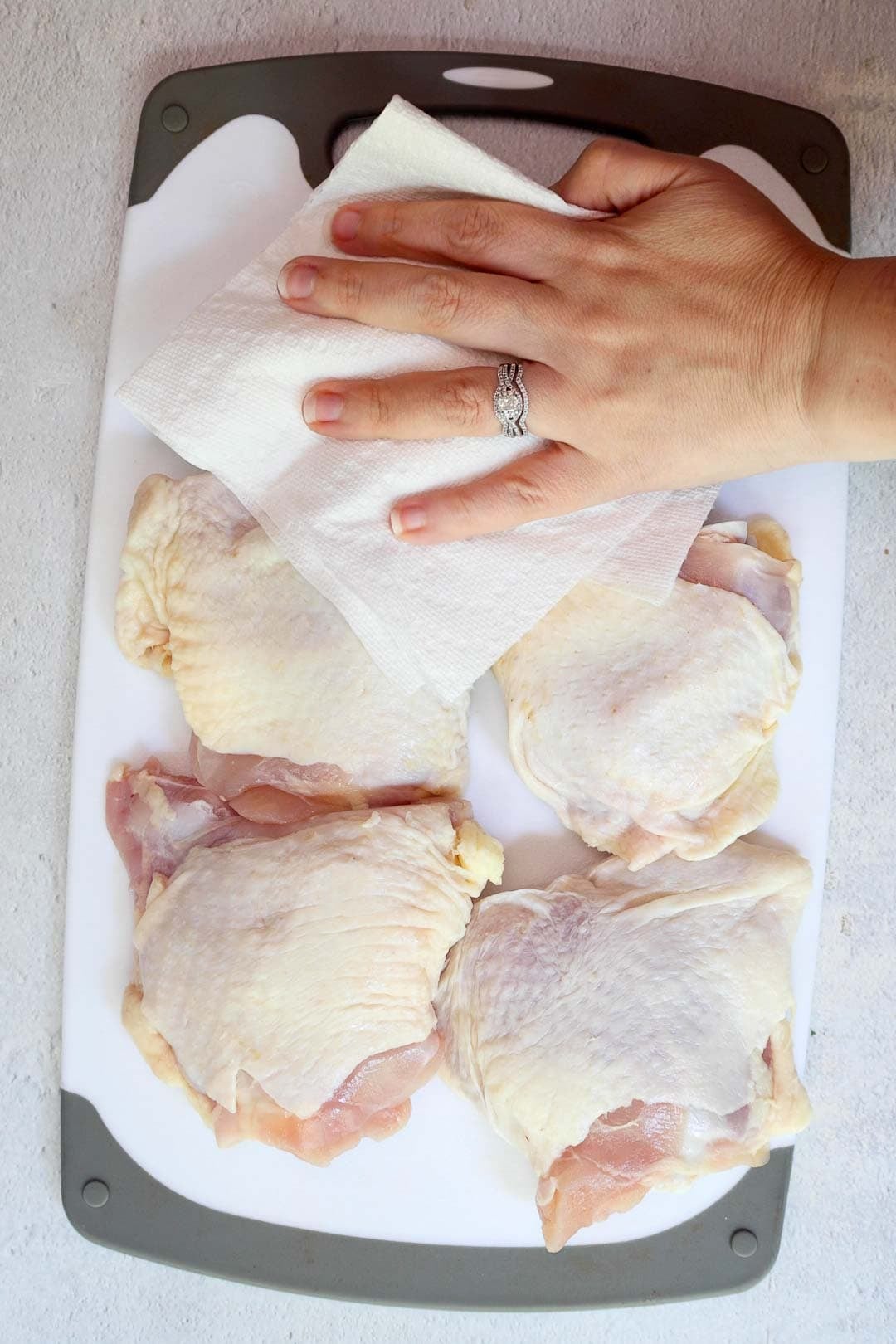 A hand using a paper towel to pat dry raw chicken thighs on a white cutting board.
