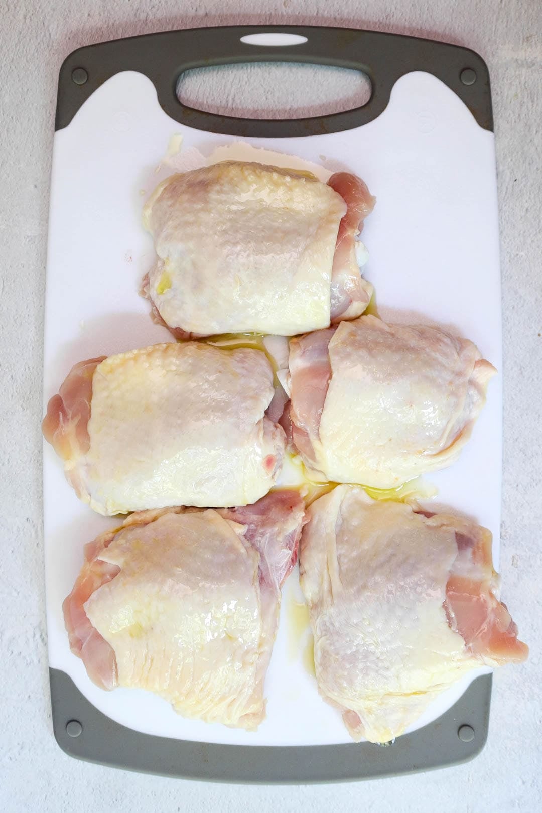 Five raw chicken thighs drizzled with olive oil placed on a cutting board.