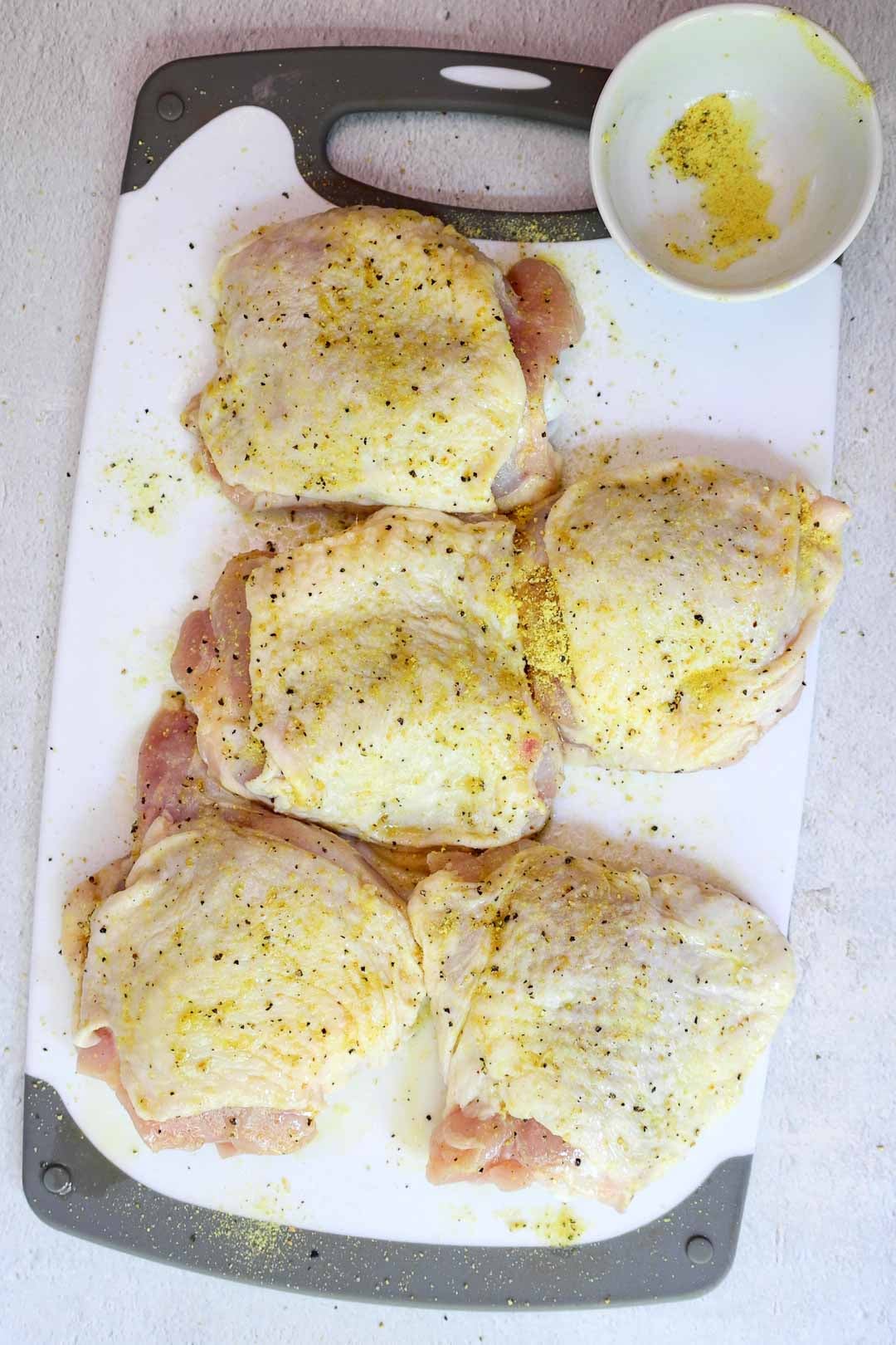 Raw chicken thighs thoroughly seasoned with lemon pepper blend on a cutting board, with visible specks of black pepper and yellow seasoning.