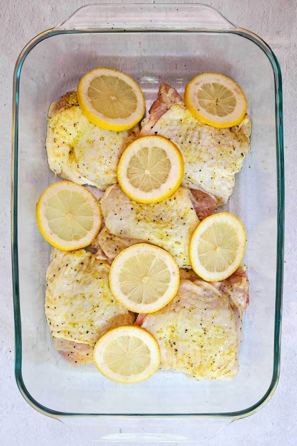 Seasoned raw chicken thighs arranged in a glass baking dish, topped with lemon slices ready for baking.
