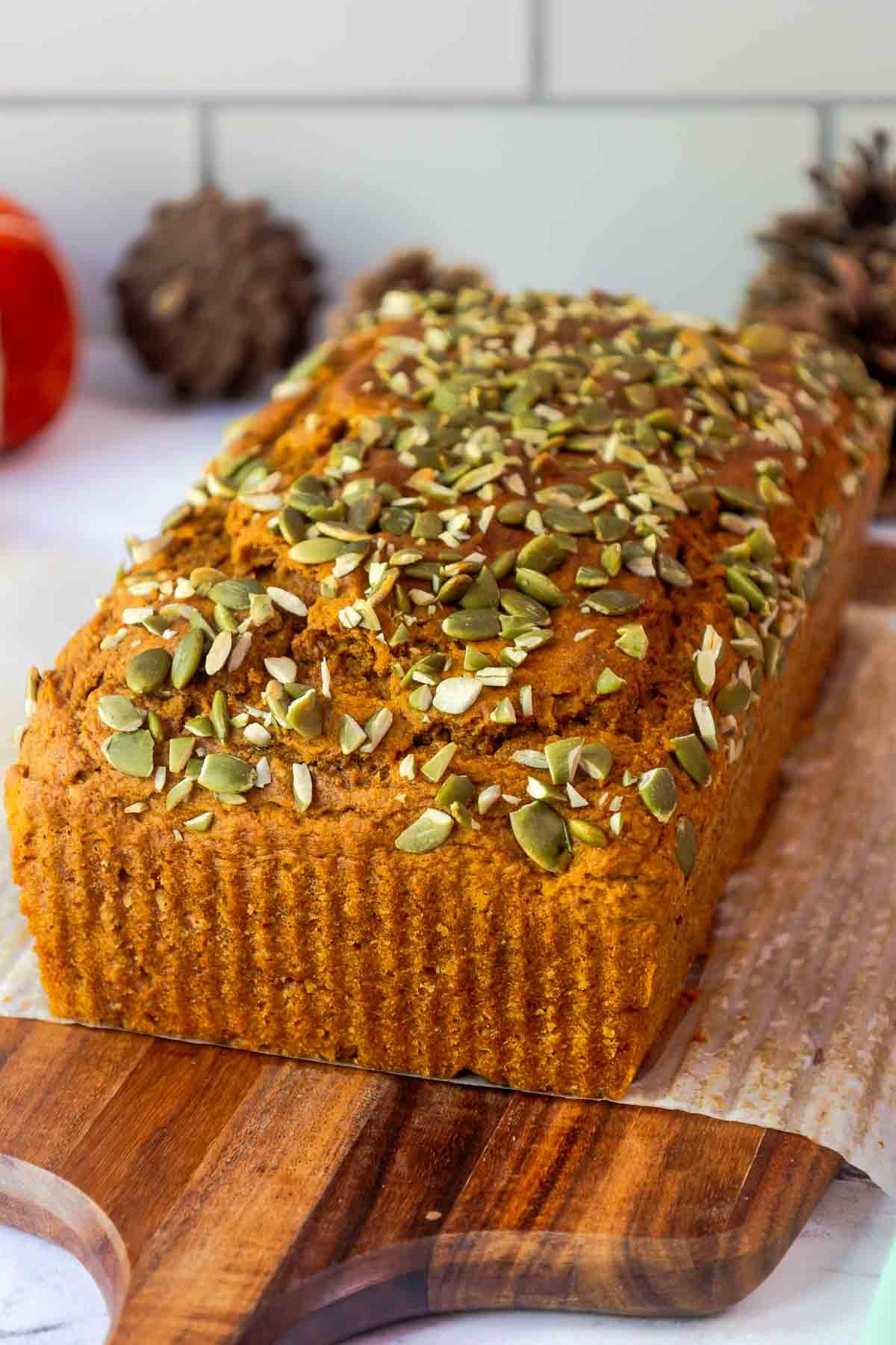 A whole loaf of golden pumpkin bread with a domed top, generously sprinkled with pumpkin seeds, resting on a wooden cutting board.
