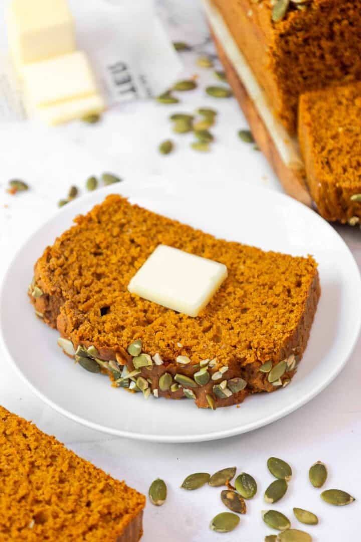 A single slice of pumpkin bread on a white plate with a pat of butter melting on top.