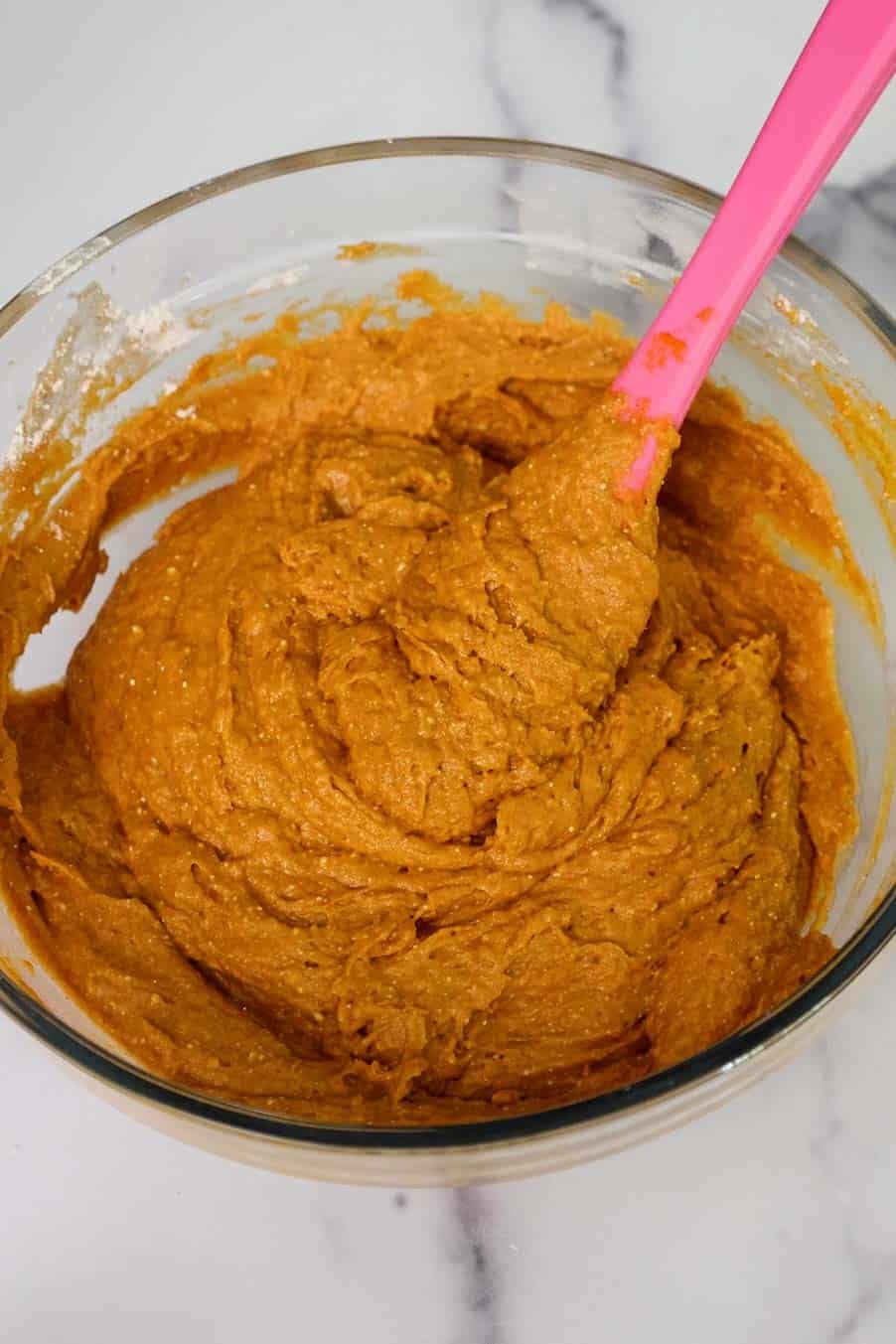 A glass mixing bowl filled with thick, pumpkin bread batter being stirred with a silicone spatula.