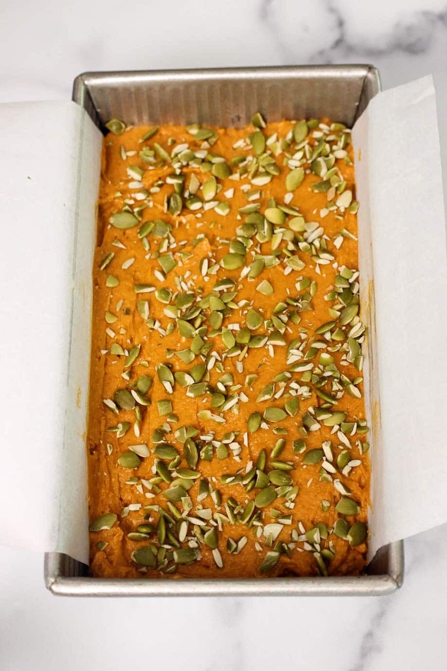 Unbaked pumpkin bread batter topped with a generous layer of pepitas, evenly spread in a parchment-lined loaf pan.