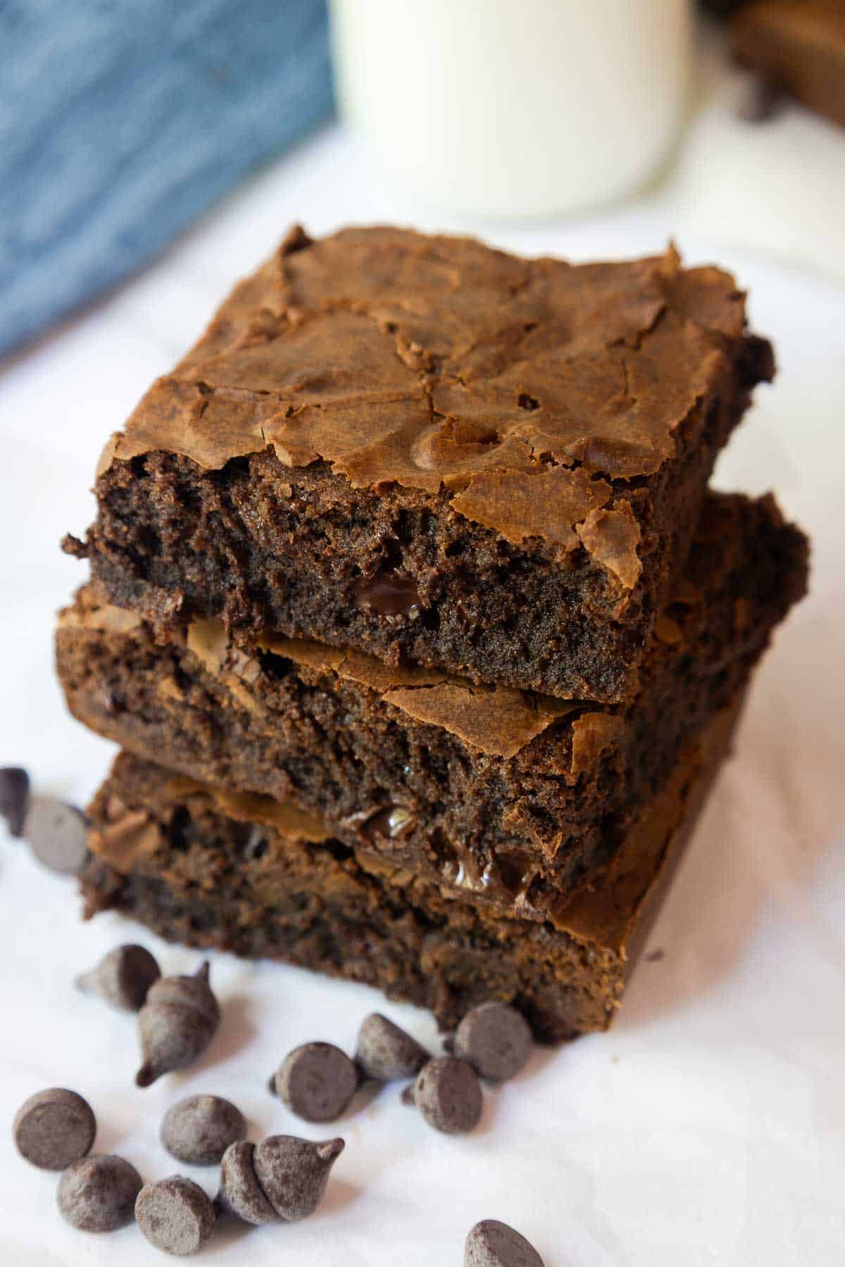 Stack of brownies next to chocolate chips and a glass of milk, highlighting the moist interior and crisp top layer.