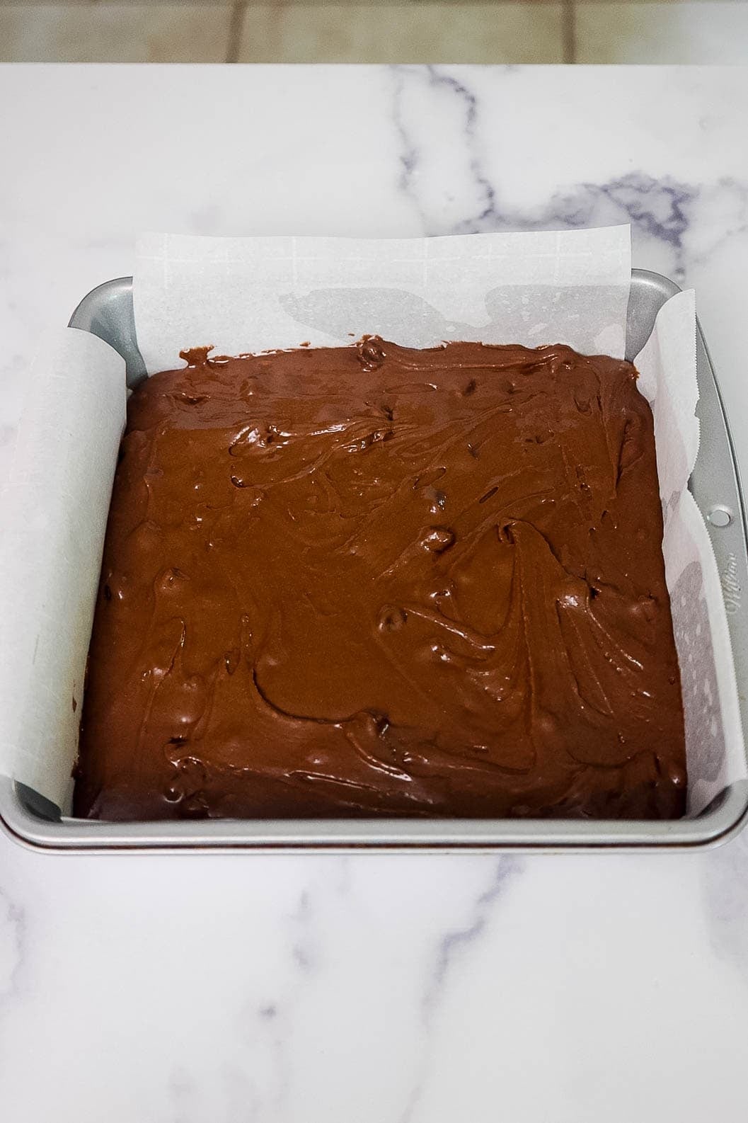 Brownie batter spread evenly in a parchment-lined square baking pan, ready to be baked.