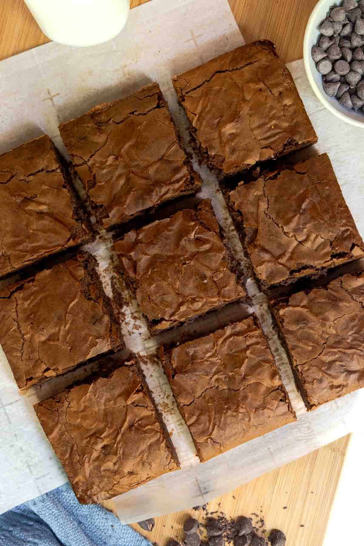 Top-down view of nine evenly cut brownies on parchment paper with a glass of milk and bowl of chocolate chips nearby.