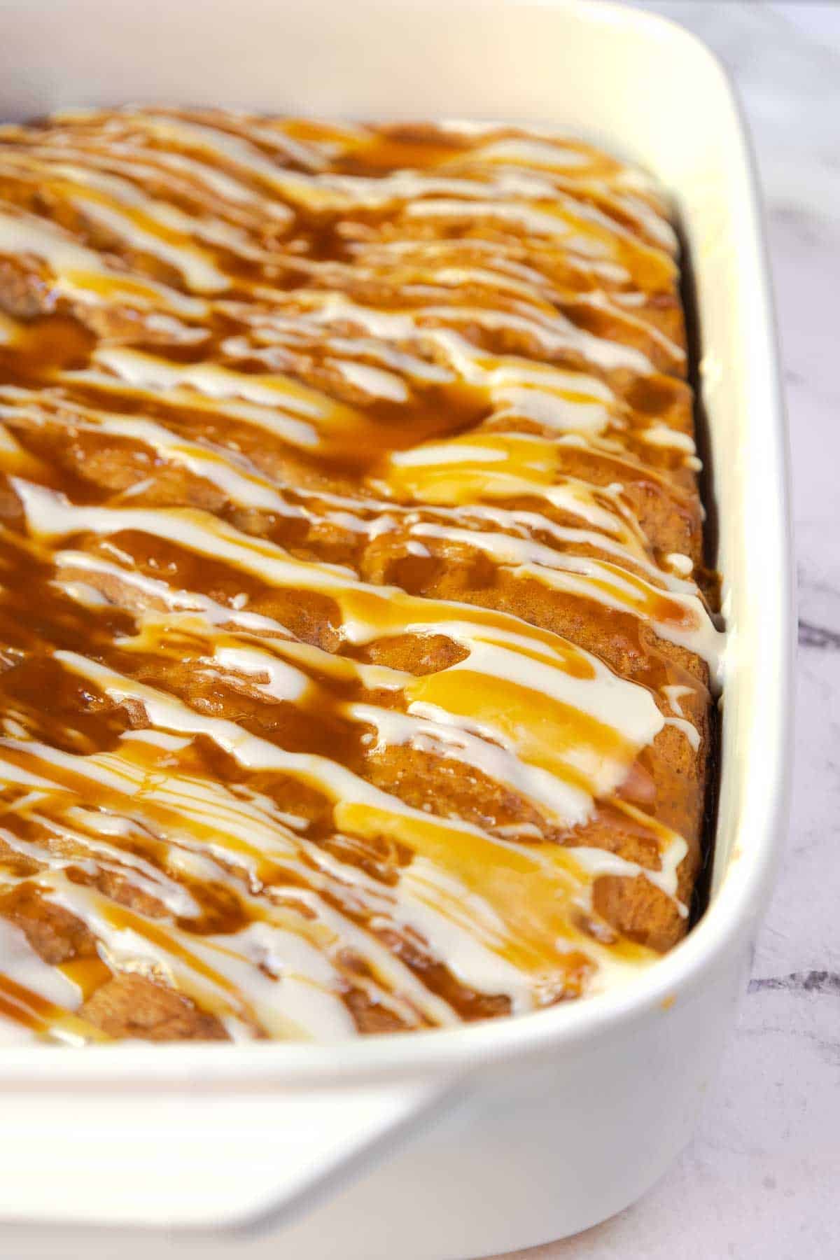 Overhead view of a golden-brown apple cake covered in zigzag drizzles of powdered sugar glaze and caramel sauce in a 9 by 13 inch baking dish.