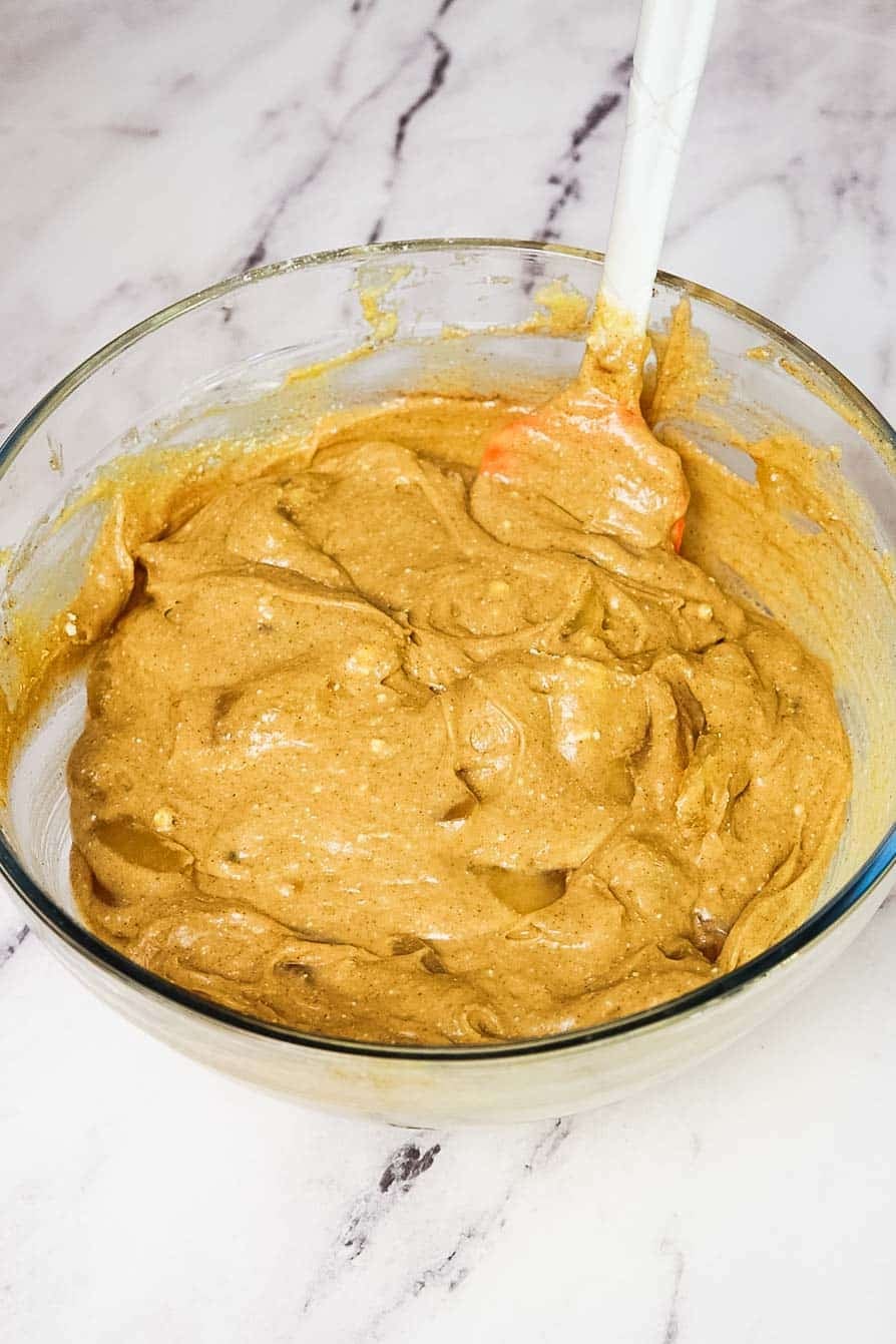Thick, cinnamon-speckled apple cake batter in a glass mixing bowl with a spatula, ready to be poured into a baking dish.